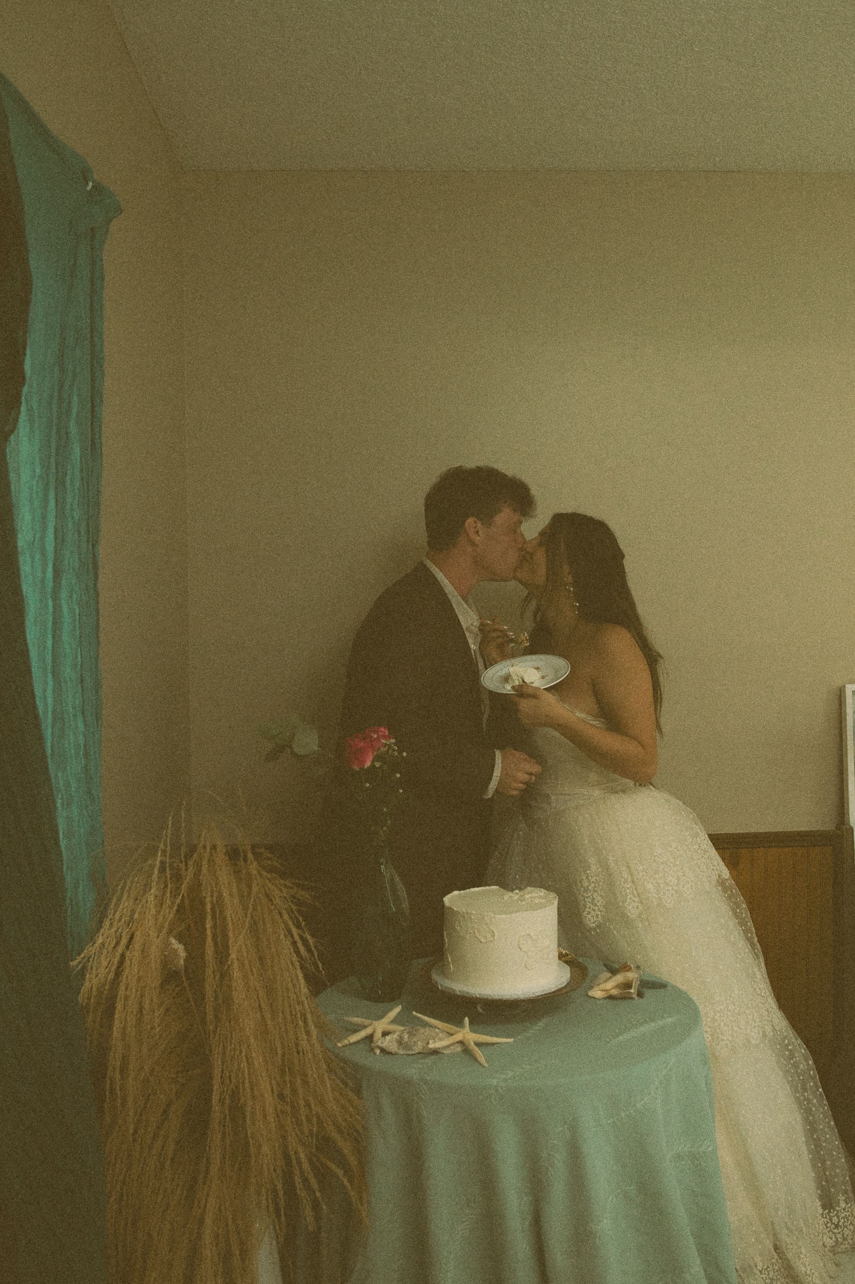 Bride and groom sharing a kiss while holding a slice of cake at an intimate reception in soft, warm light.