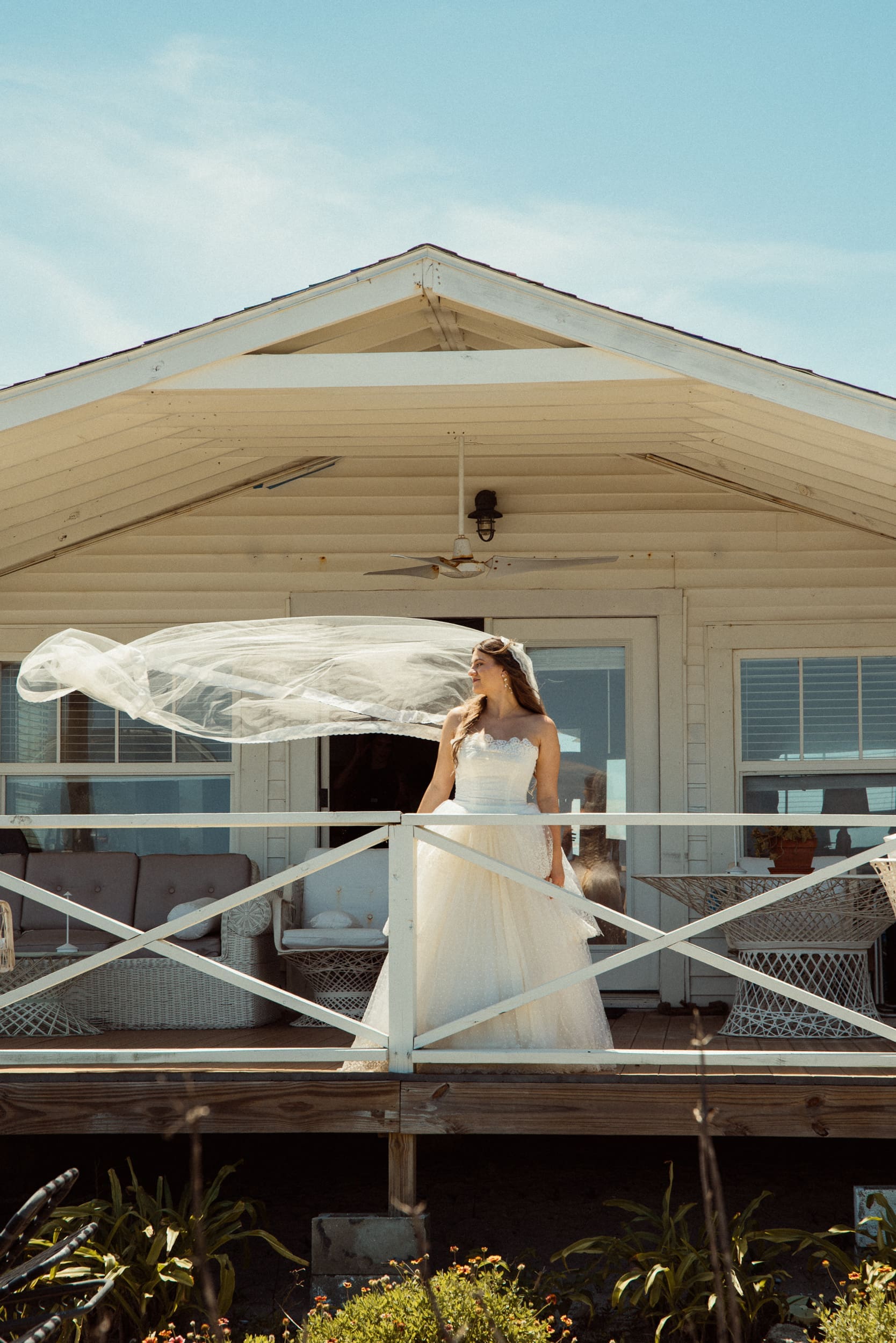 Bride on a white porch as her long veil lifts in the wind, an editorial moment shaped around natural movement.