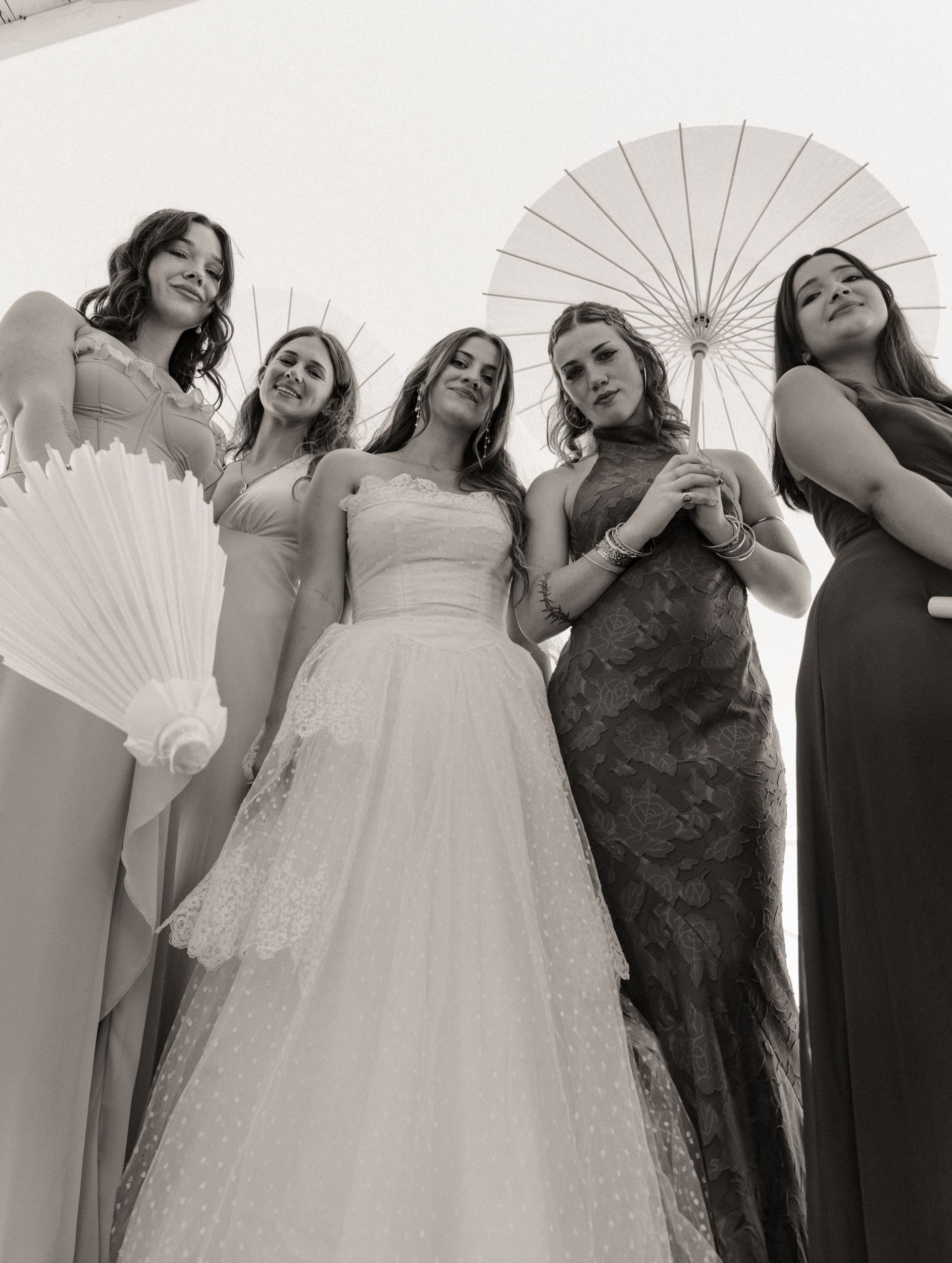 Low-angle black and white portrait of a bride and bridesmaids with a paper parasol, a playful editorial wedding party photo.