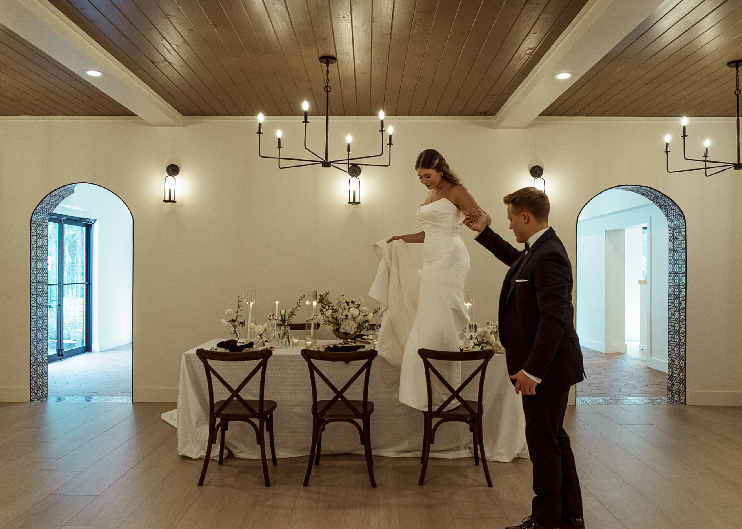 Editorial wedding portrait of a bride stepping onto a reception table with her groom's support, symbolizing trust, movement, and effortless artistry in wedding photography.