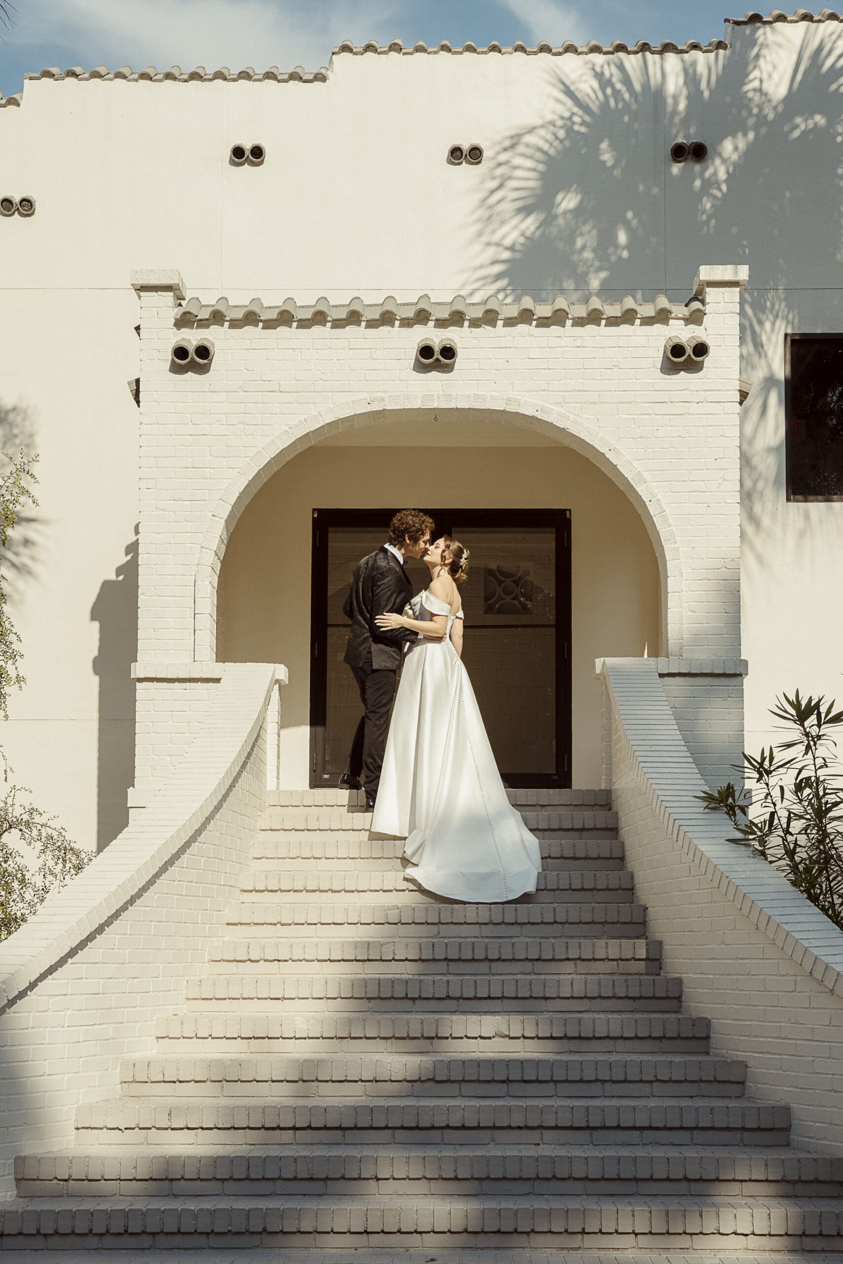 Editorial wedding portrait of bride and groom sharing a kiss on sunlit white steps, captured in a timeless, cinematic composition. 