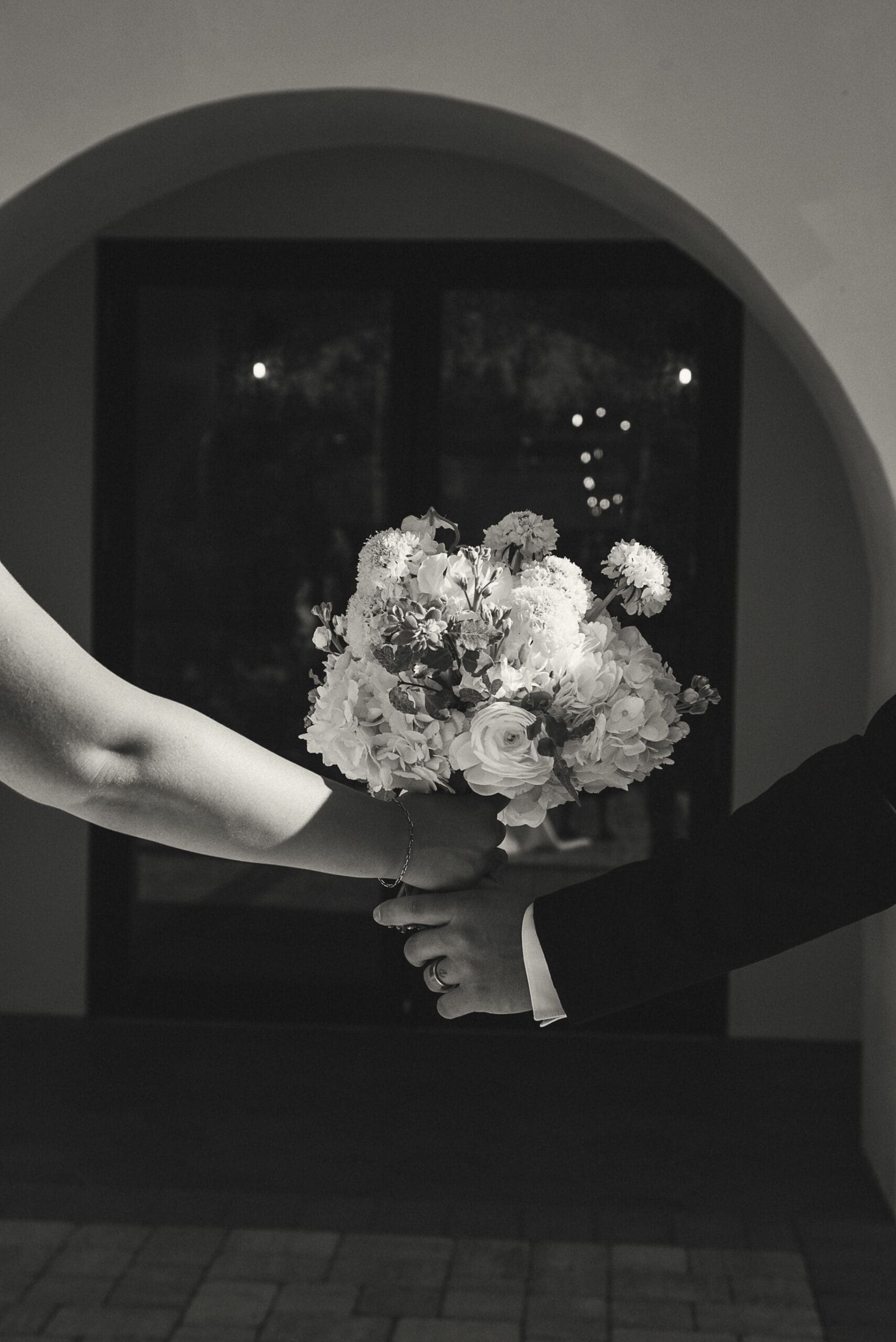 Black and white editorial shot of a bride and groom's hands holding a bouquet, symbolizing unity and simplicity in timeless wedding imagery. 