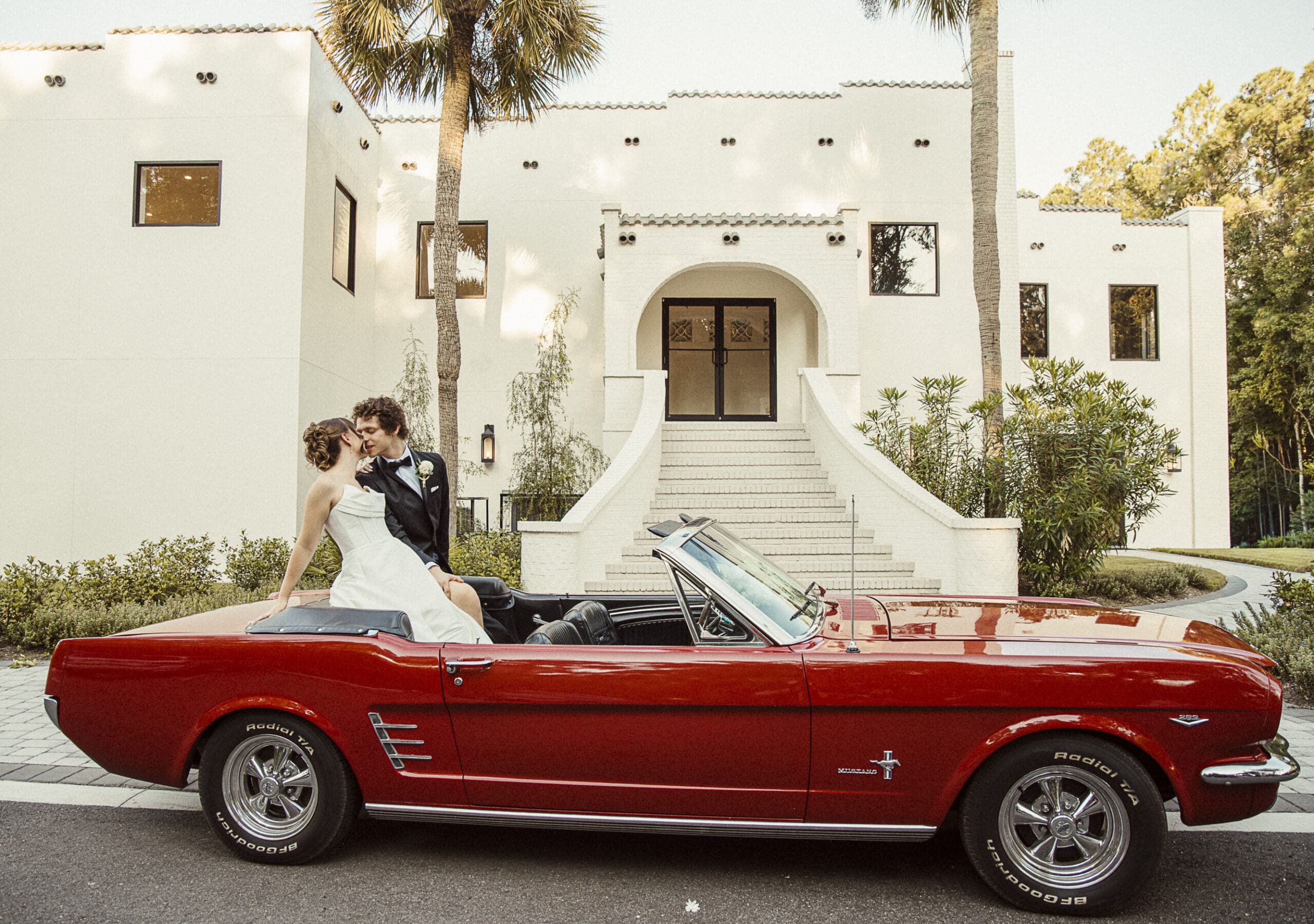 Editorial wedding portrait of a bride and groom sharing a kiss in a vintage red convertible outside a white art-deco building in Jacksonville, Florida, captured with timeless, cinematic style.