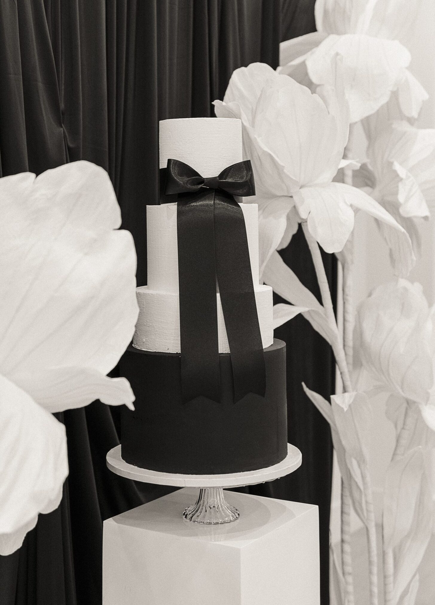 Black and white tiered wedding cake with an oversized ribbon bow, framed by sculptural white florals.
