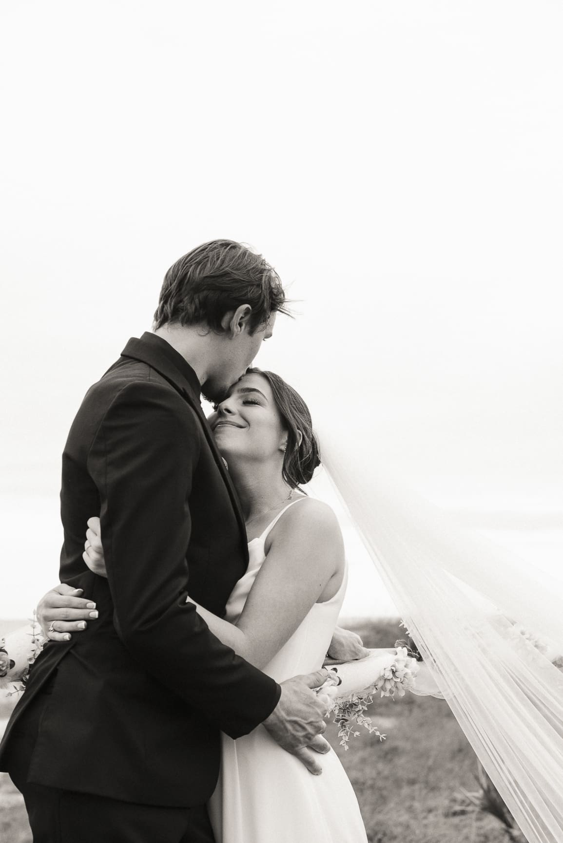 Intimate editorial wedding portrait of groom kissing his bide's forehead, capturing calm emotion and effortless connection in timeless style.