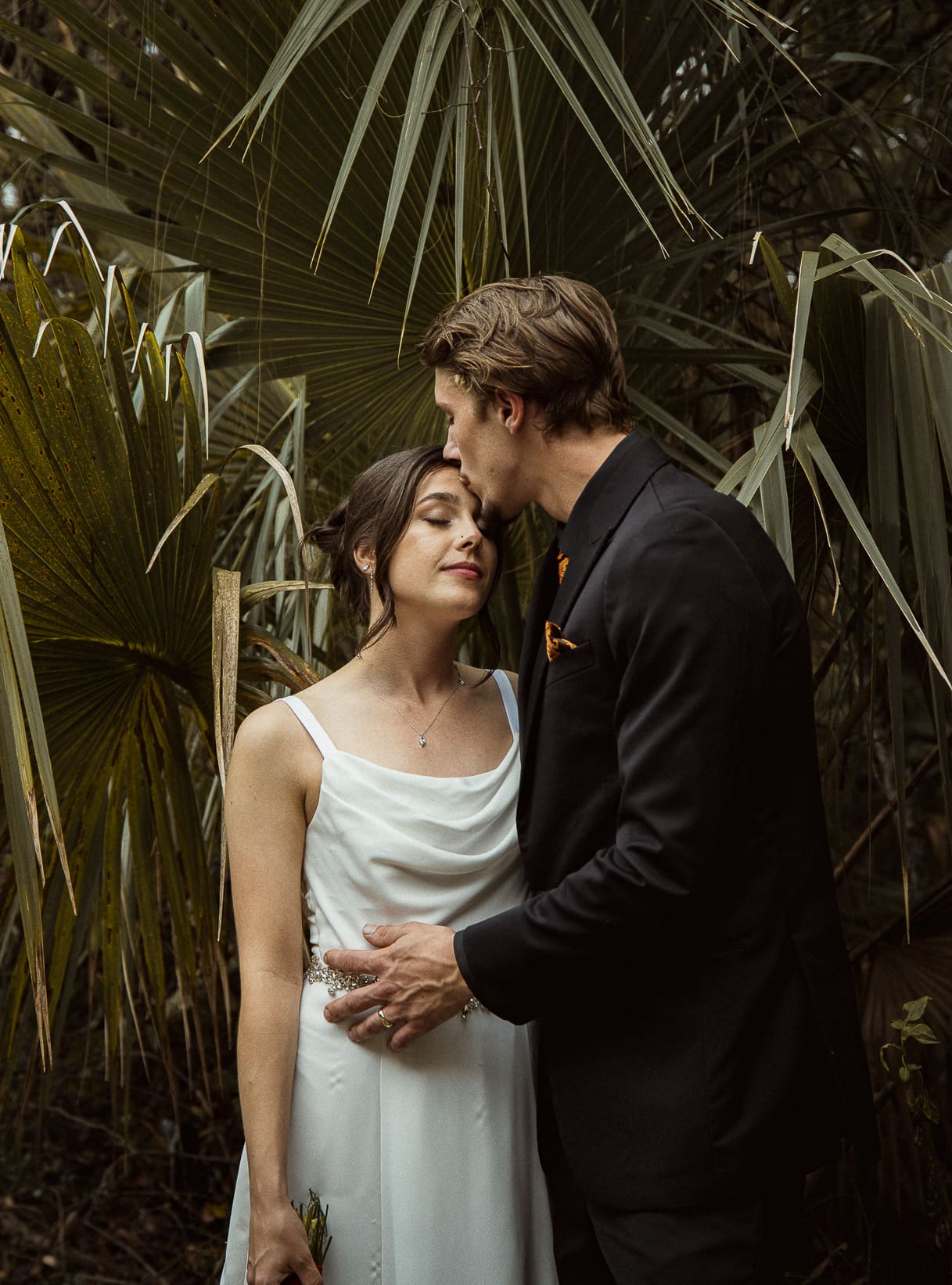 Editorial wedding portrait of a groom kissing his bride's forehead surrounded by tropical palms, representing intimacy and ease through effortless wedding photography. 