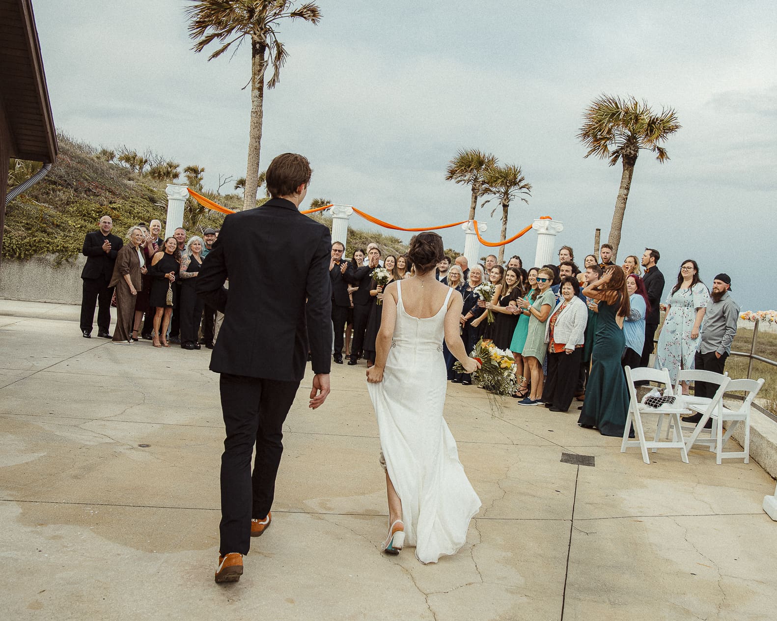 Editorial wedding moment as couple walks hand-in-hand toward celebrating guests, showcasing joy and movement through cinematic composition.
