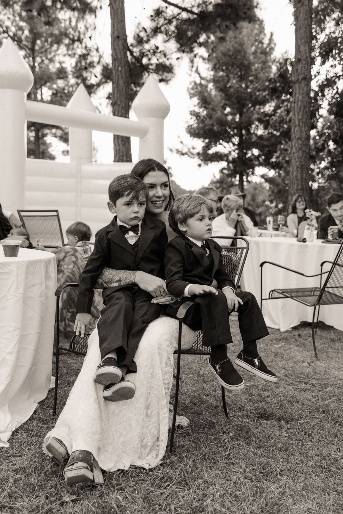 Bride in her wedding dress holds two young ring bearers in suits on her lap at an outdoor reception