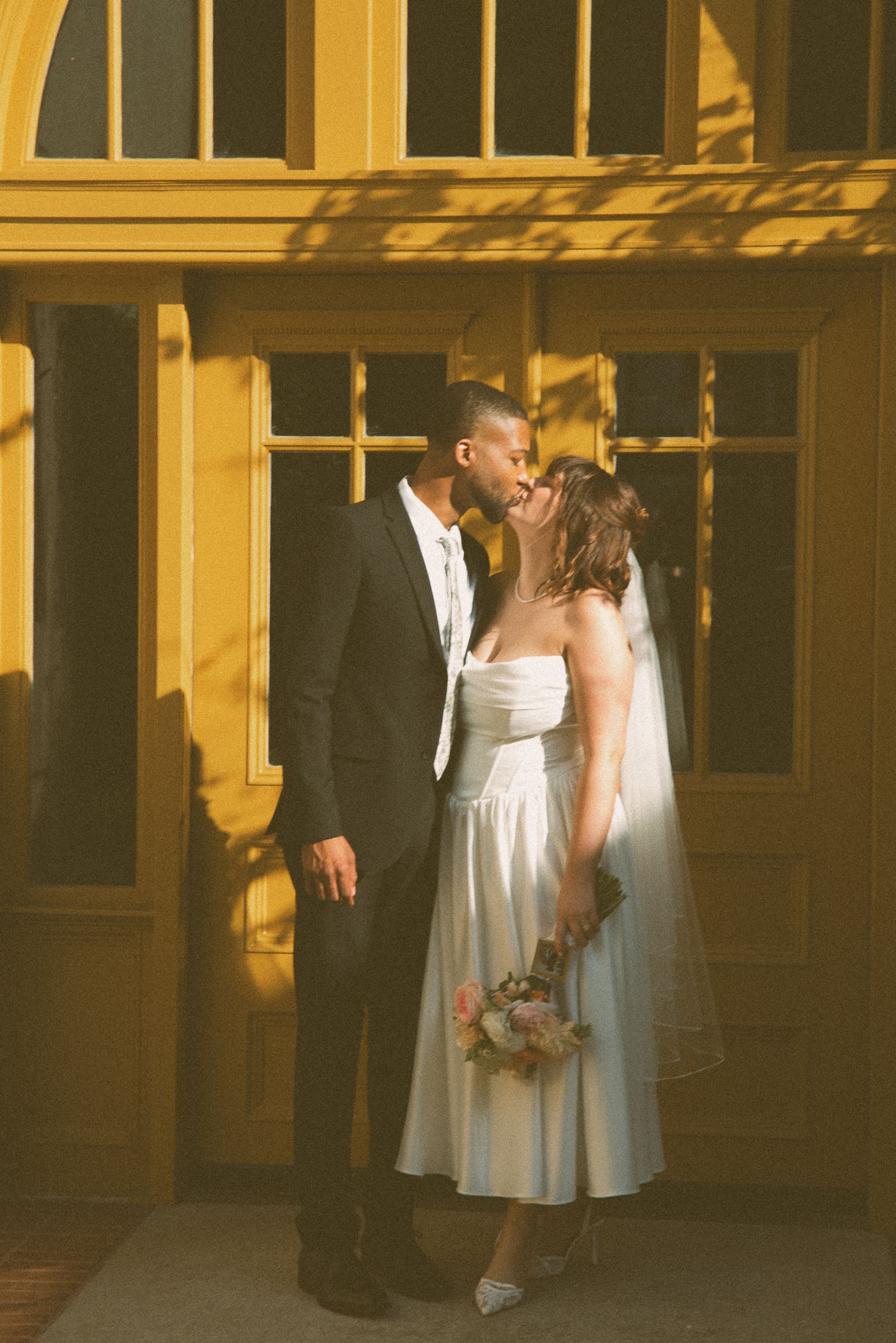 Bride and groom kissing in low golden light against a historic yellow door in downtown St. Augustine dappled with leaf shadows.