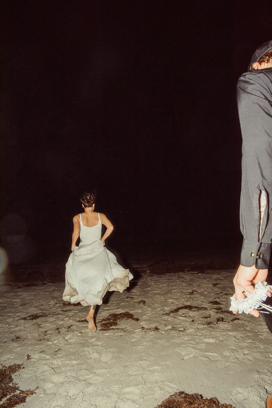 Bride running barefoot across the sand at night on Jacksonville beach, lit by flash, as the groom looks on. A spontaneous editorial wedding moment.