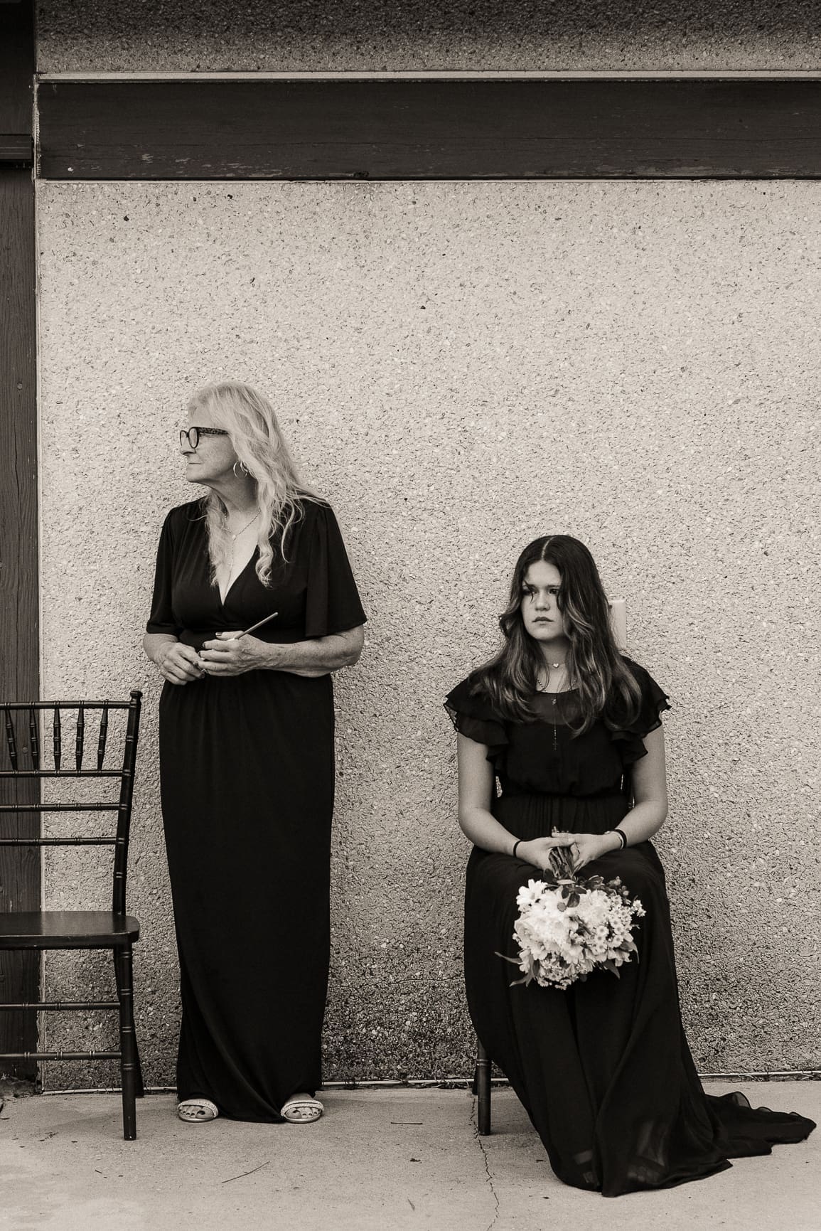 Black and white editorial portrait of two women in black dresses against a textured wall, one seated holding a bouquet.