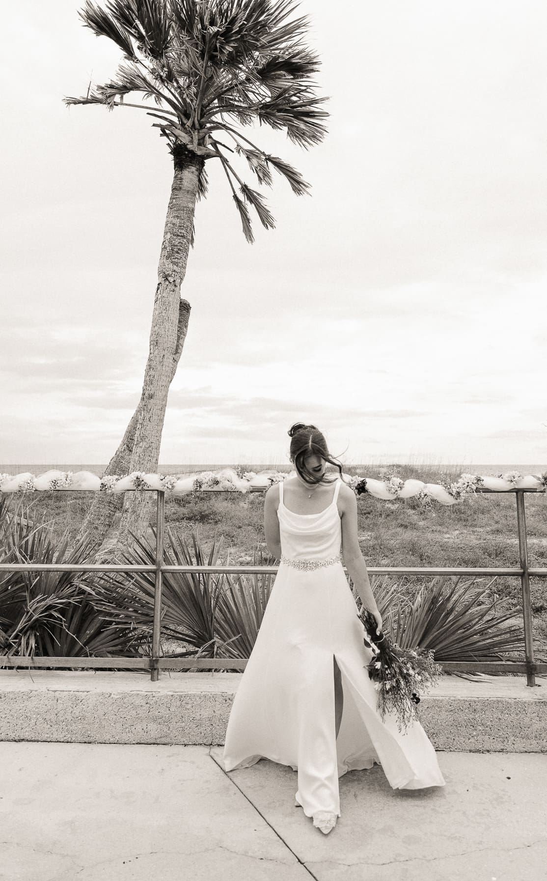 Black and white portrait of a bride walking along an oceanfront railing beneath a wind-swept palm tree.