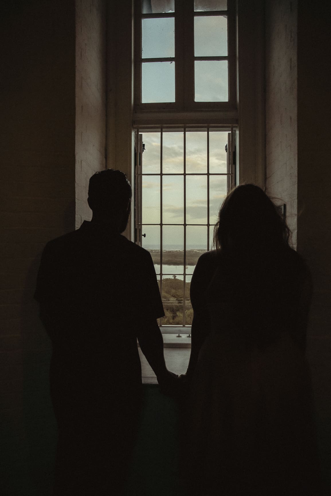 Silhouetted couple holding hands in front of a window, captured during a quiet moment created by a spacious wedding day timeline. 