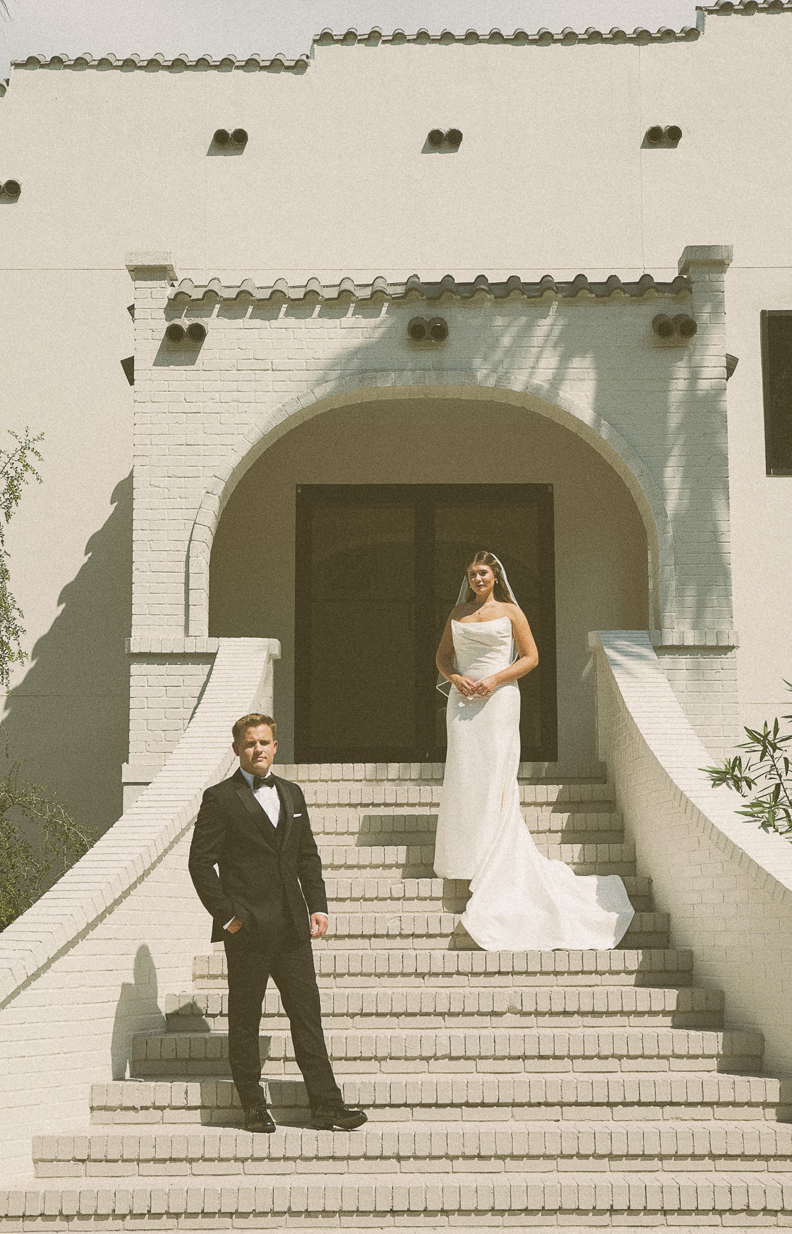 Editorial wedding portrait of a bride at the top of a Spanish-style staircase with the groom in a tuxedo standing below.