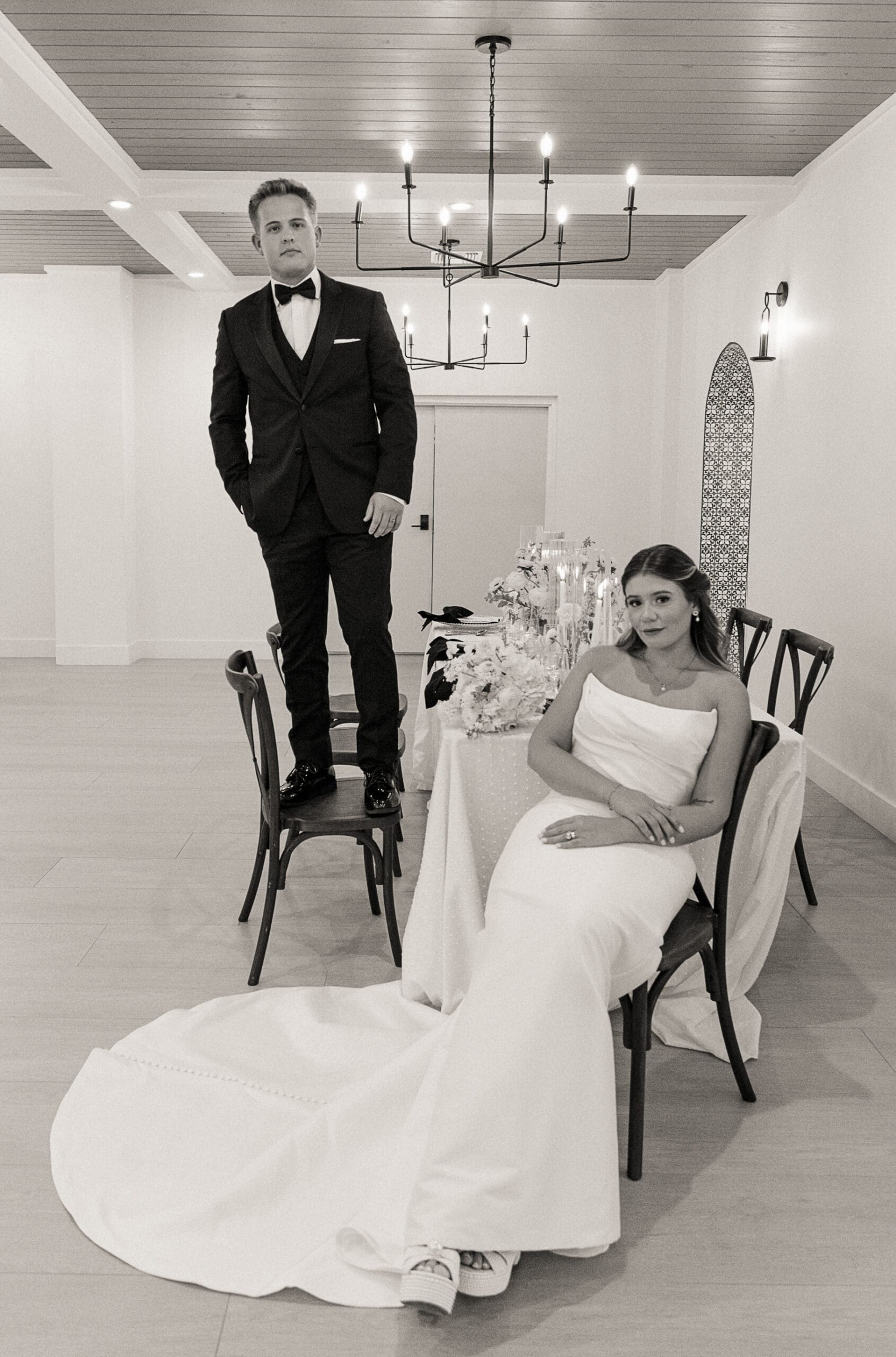 Editorial-style black-and-white portrait with the groom standing on a chair beside the seated bride