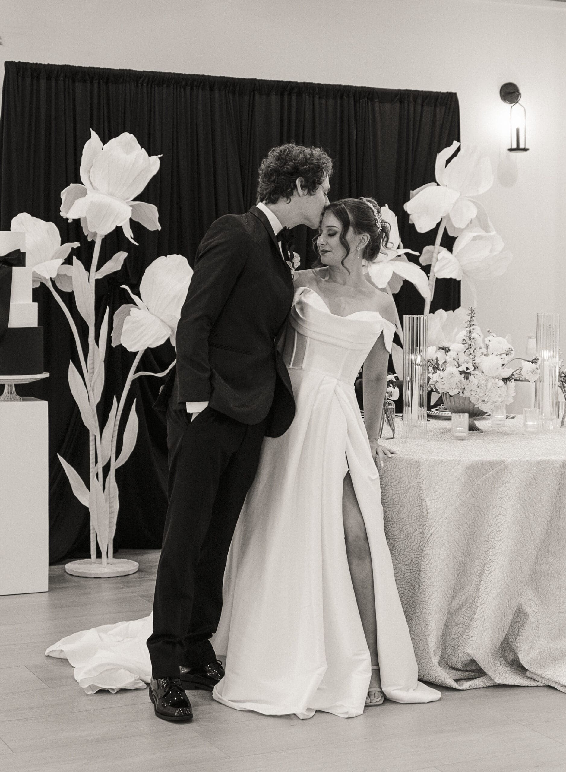 Bride and groom sharing a kiss beside the reception tablescape and paper flower backdrop