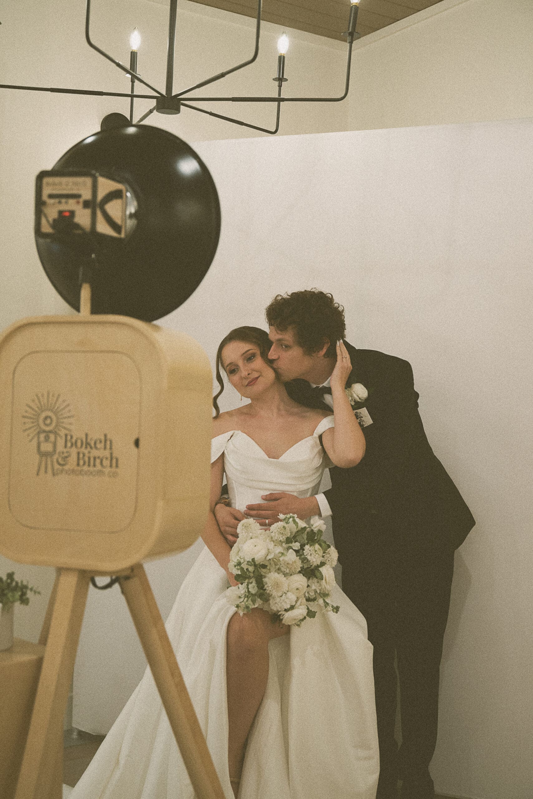Candid kiss between bride and groom in front of the Bokeh and Birch photo booth backdrop