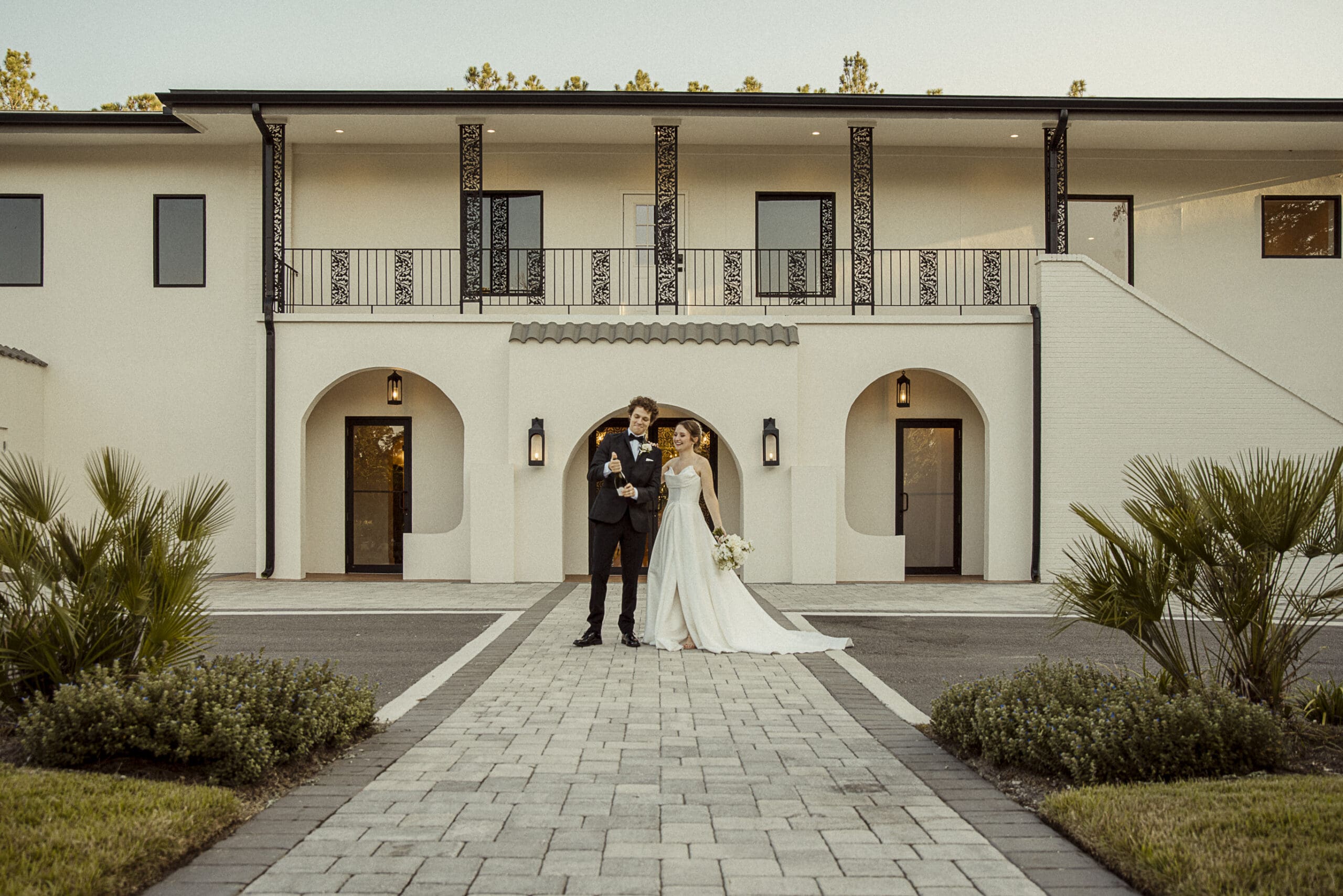 Full-length portrait of the bride and groom beneath the arched colonnade at dusk