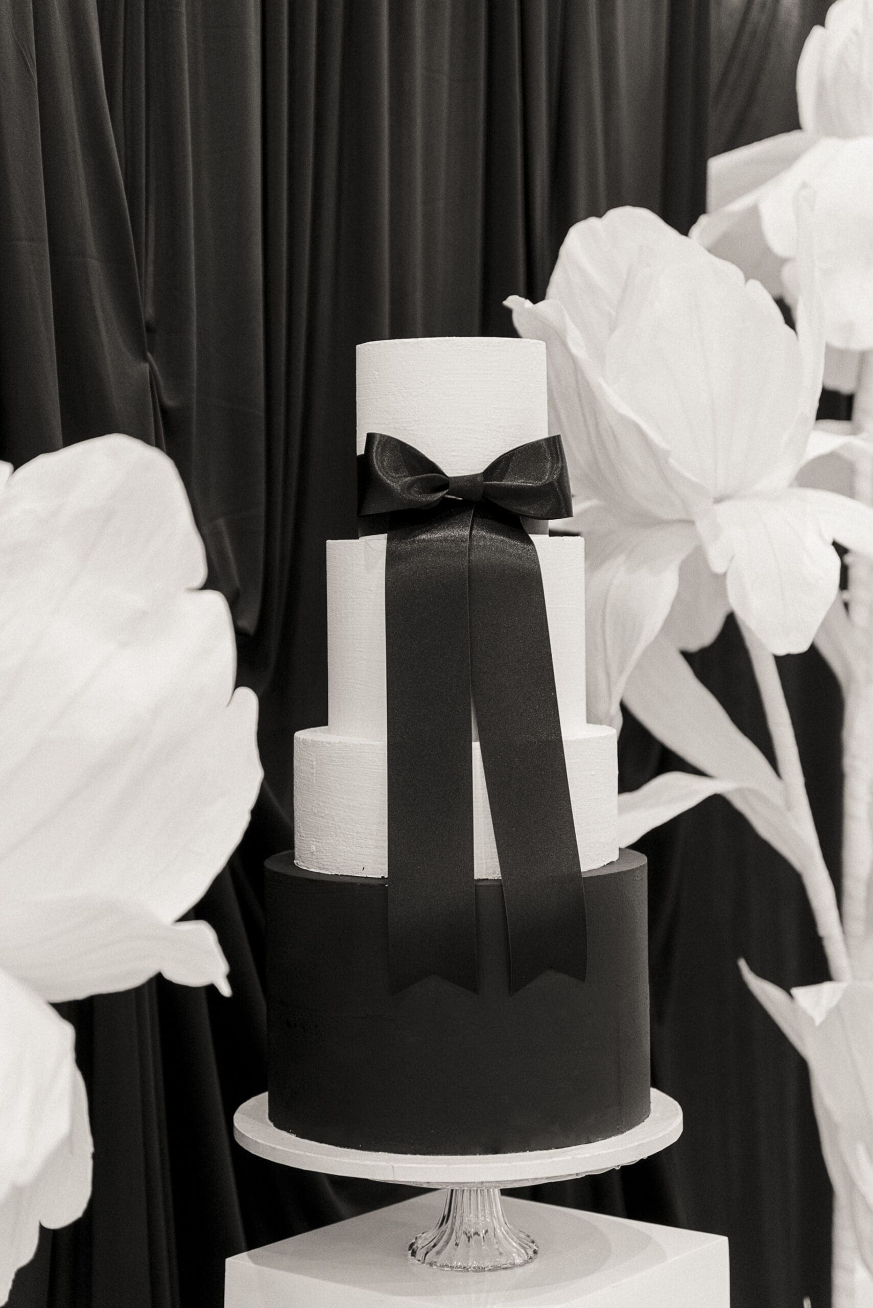 Black and white tiered wedding cake wrapped in an oversized satin bow, framed by large white paper flowers