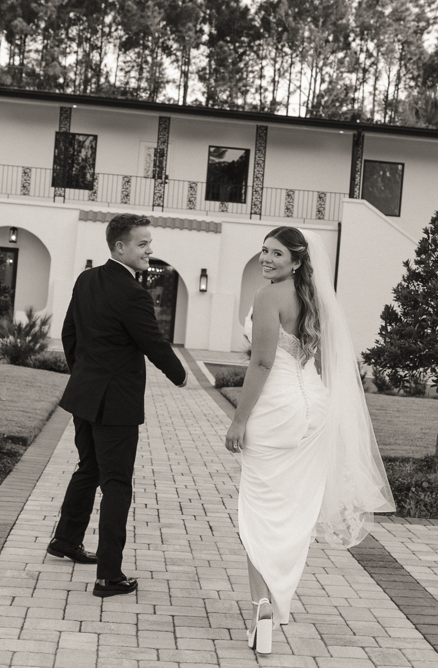 Bride glancing back at the groom while walking along the courtyard pathway, black-and-white
