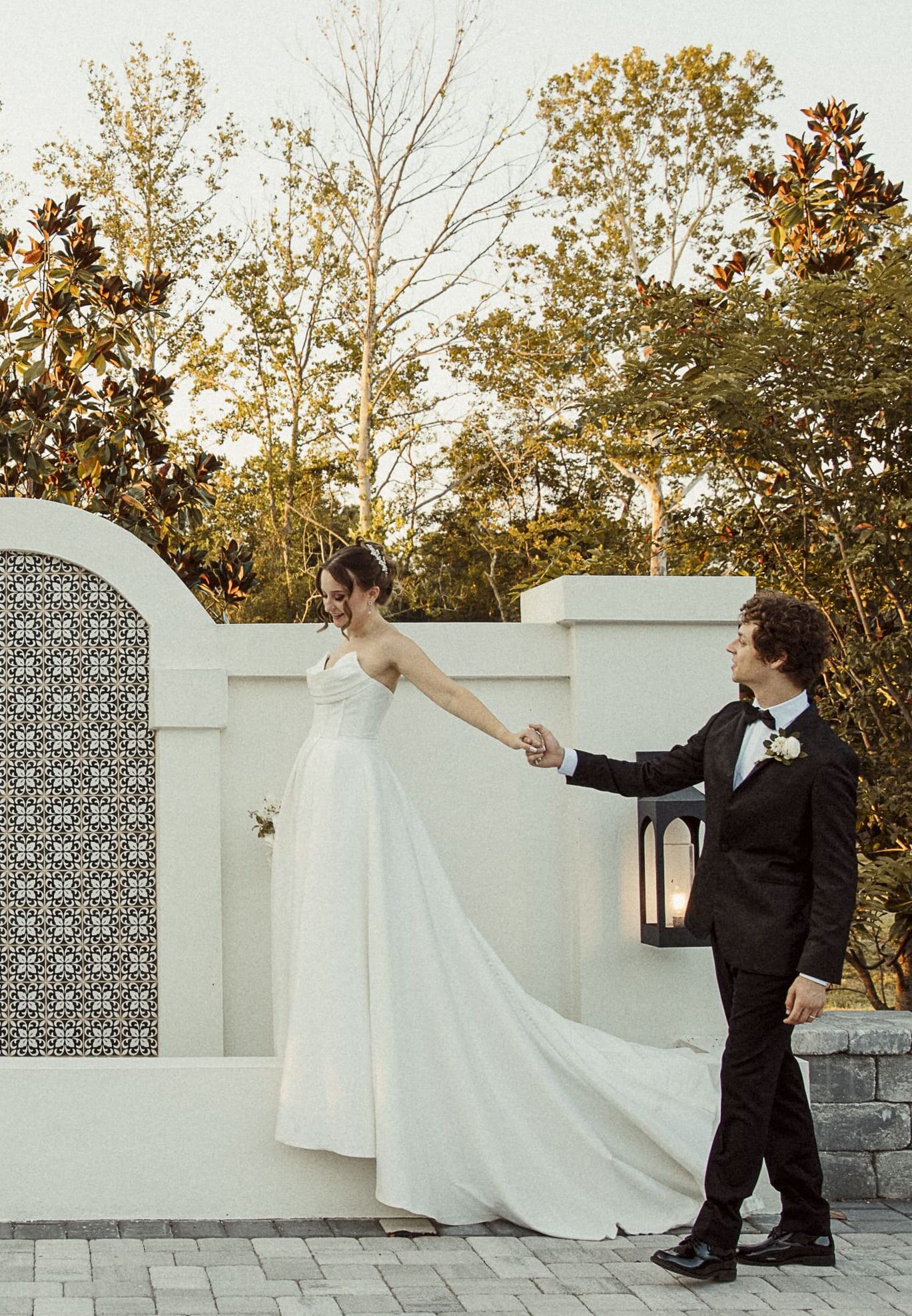Bride and groom holding hands beside a garden wall at sunset