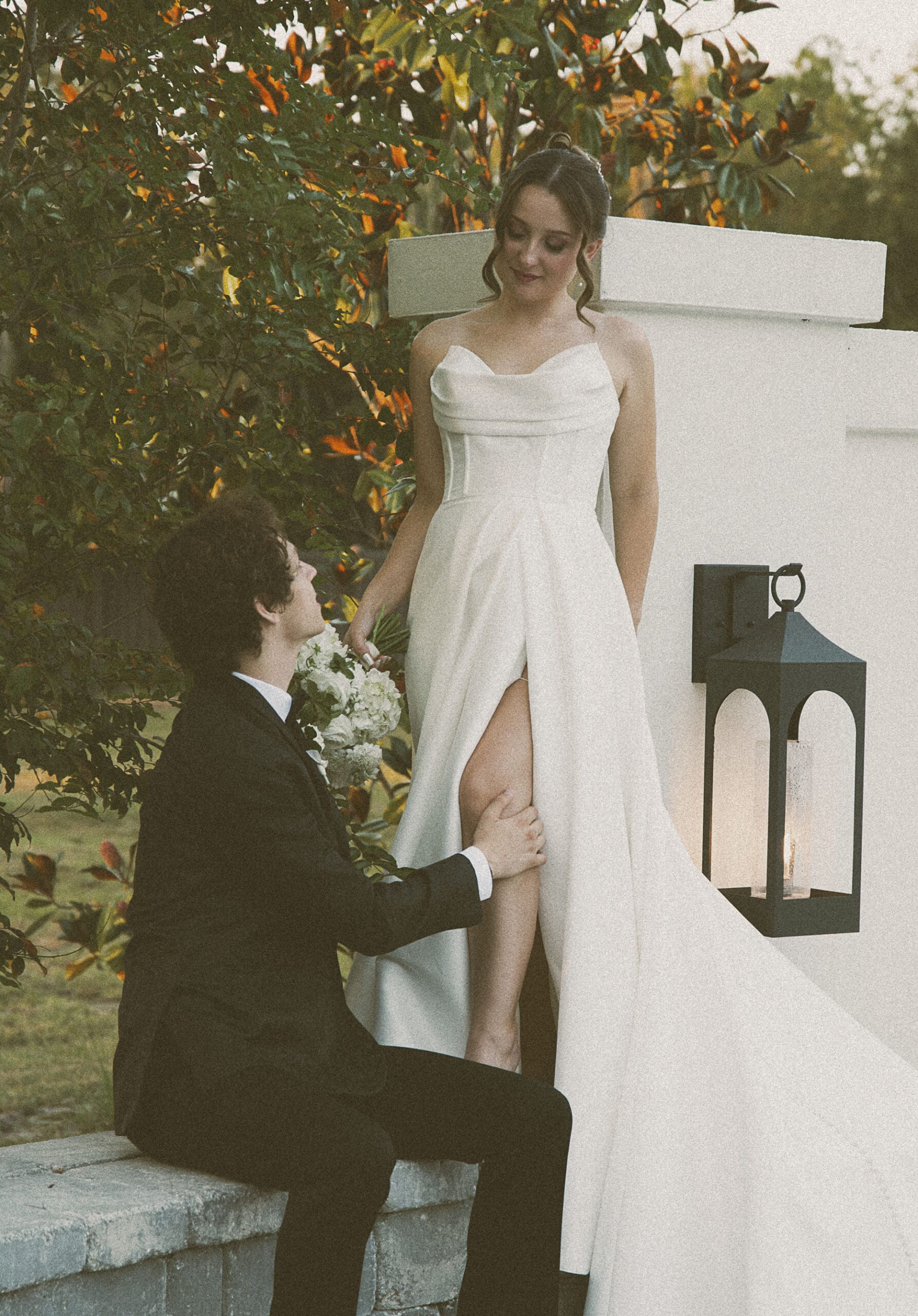 Groom helping the bride over a low courtyard wall, candid golden-hour portrait