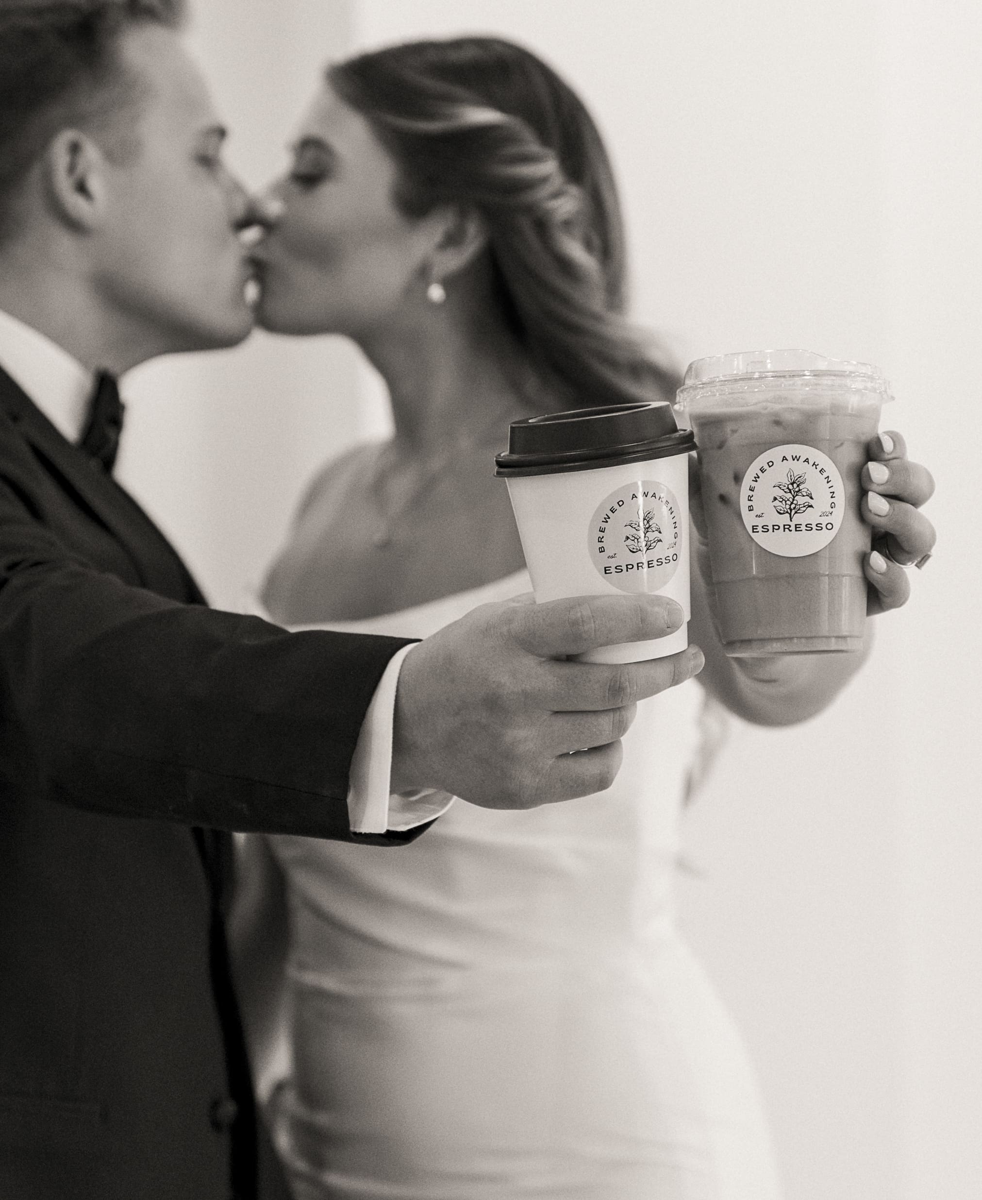 Black-and-white candid of the couple sharing a kiss while holding Brewed Awakening coffee cups