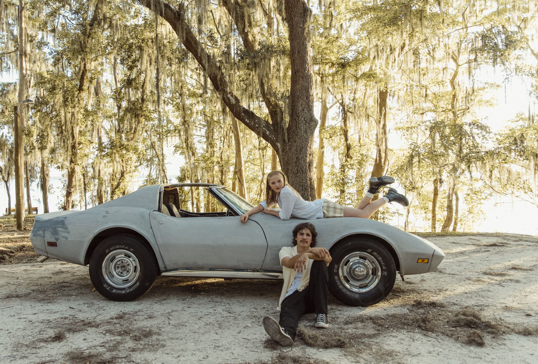 Soft-focus editorial couples portrait with woman lying on a vintage Corvette and her partner seated in front, photographed under mossy oak trees. 