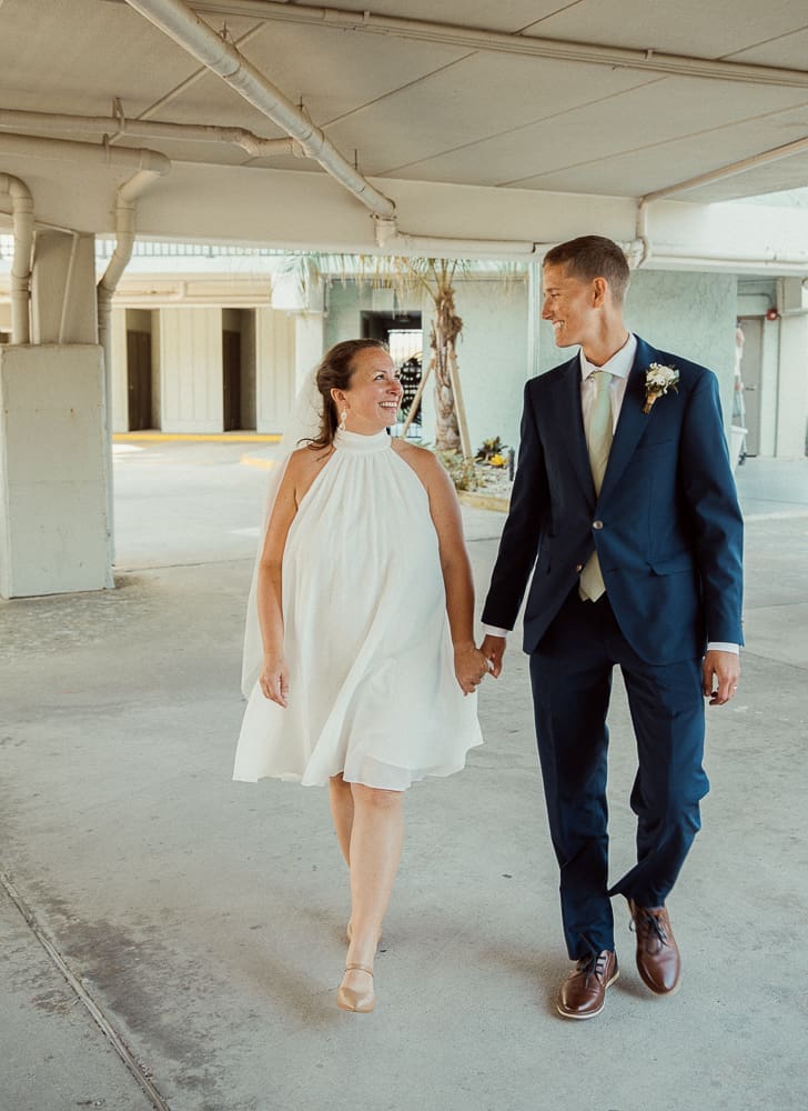Bride in a short white halter dress and groom in a navy suit walk hand in hand, smiling at each other