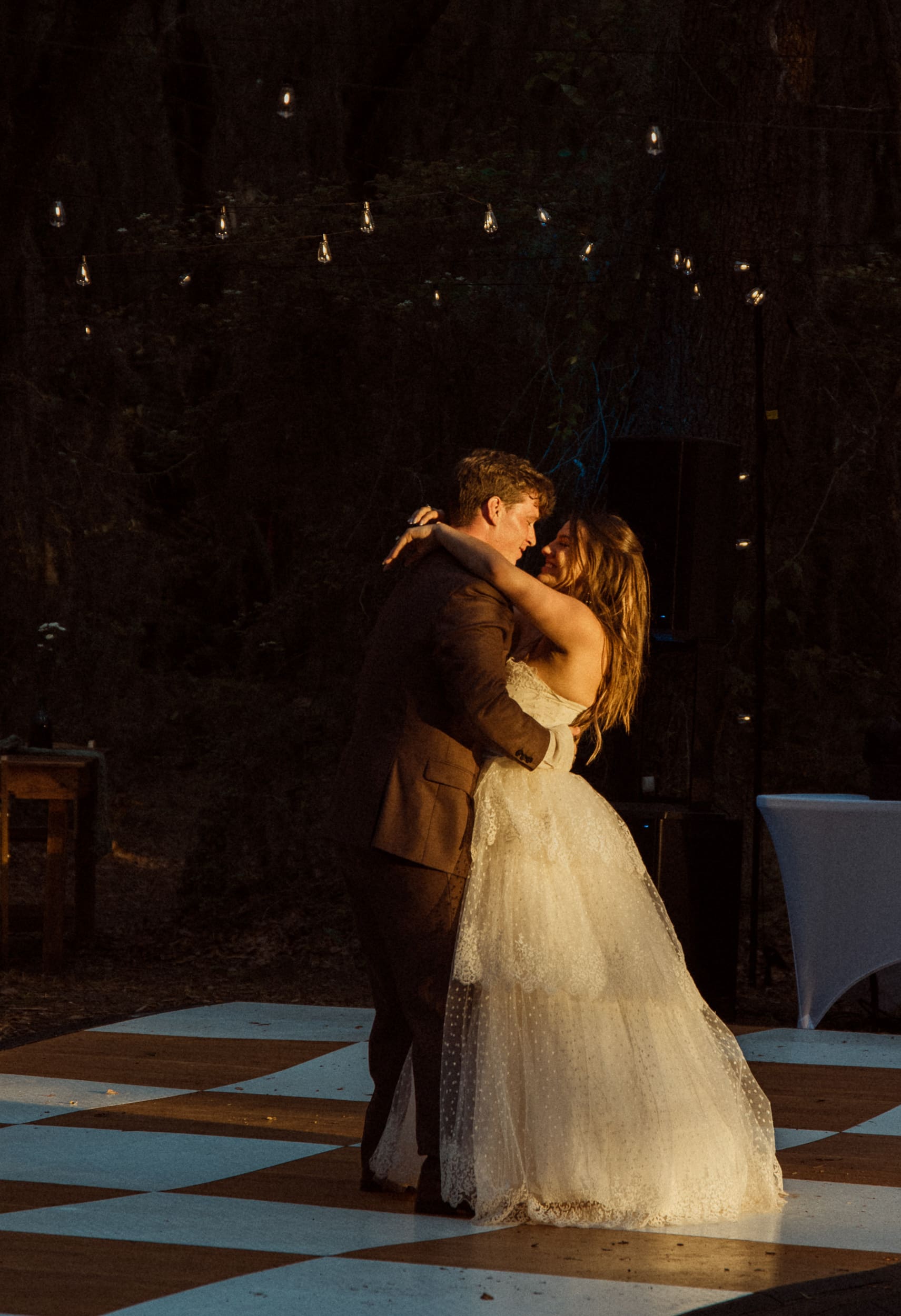 Couple shares their first dance on a checkered dance floor beneath string lights at night