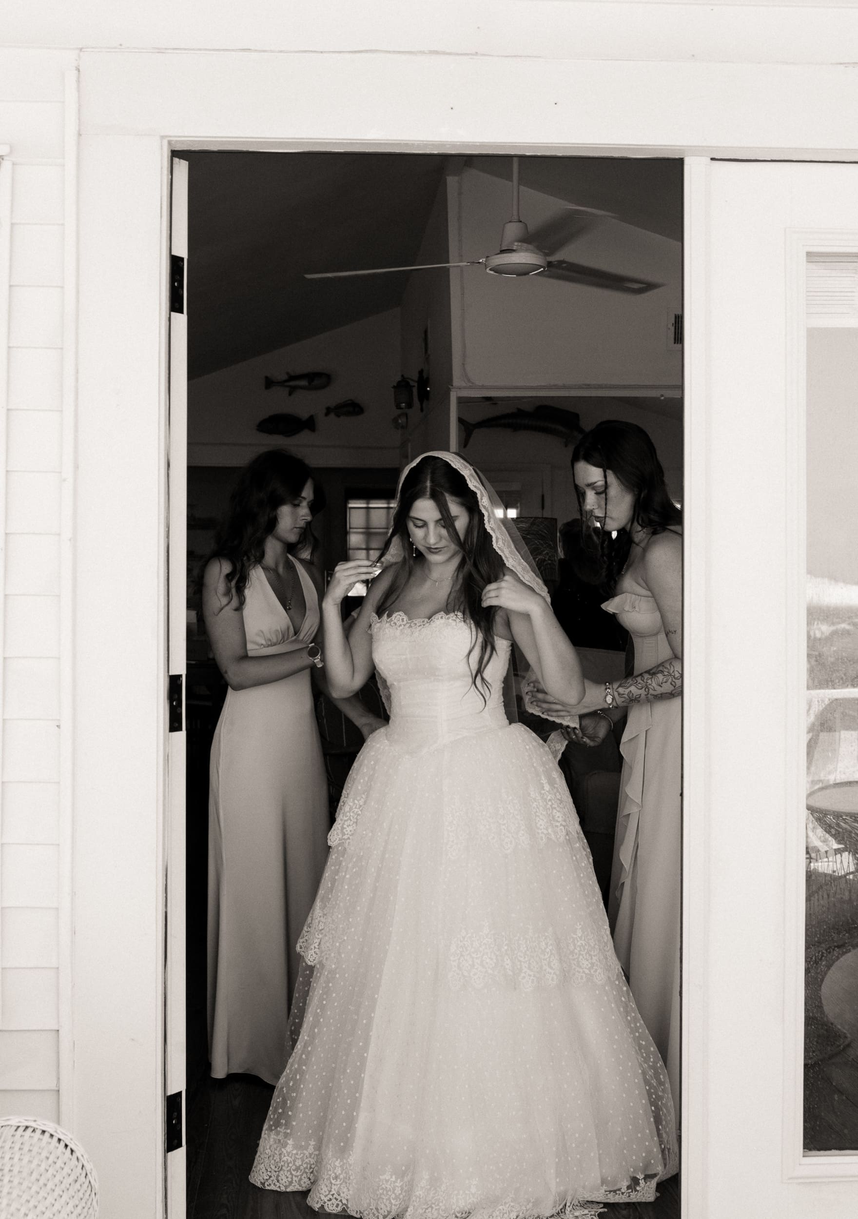 Two bridesmaids help a bride into her lace wedding gown and veil in a calm getting-ready room