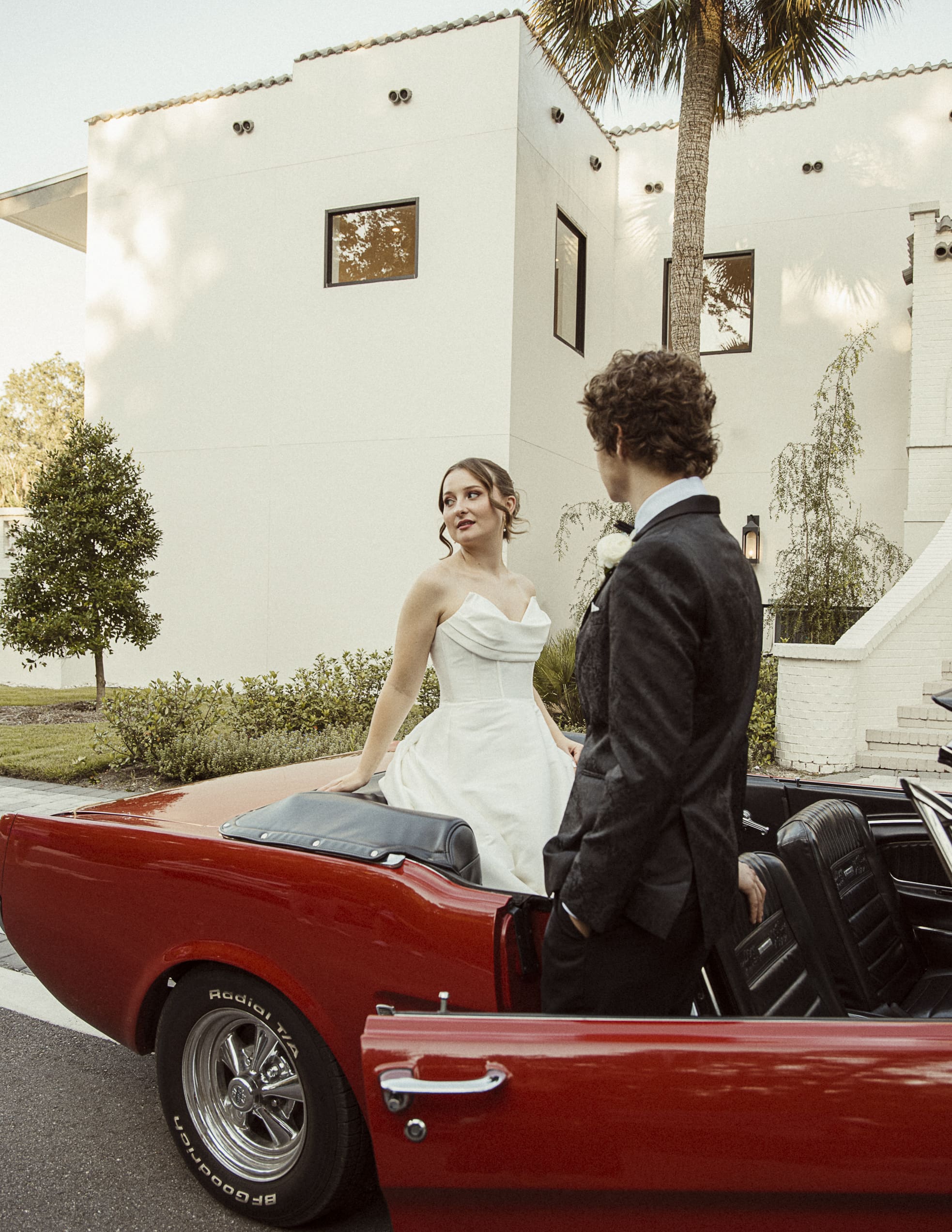 Bride and groom stand in a vintage red convertible outside a home with a palm tree