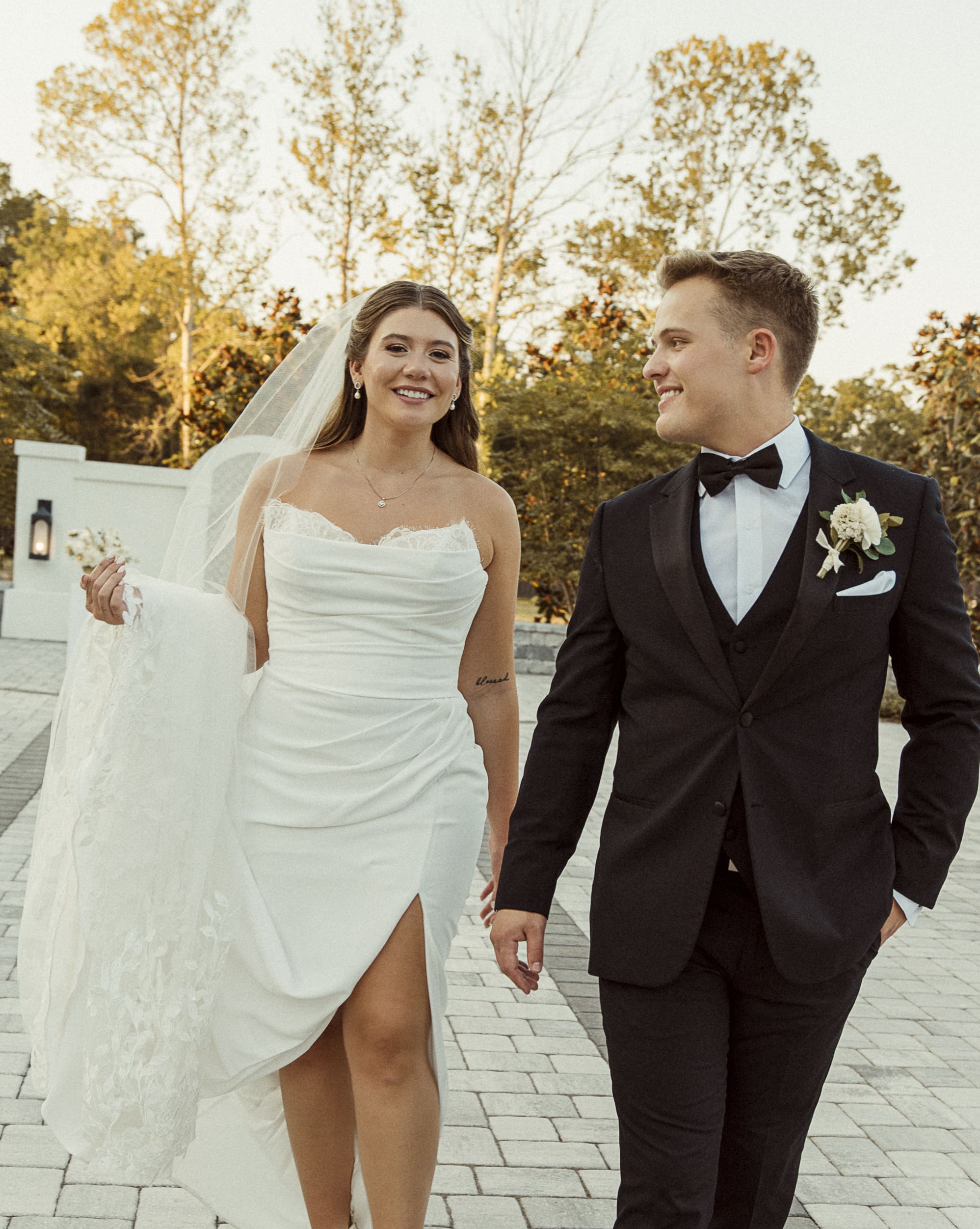 Bride in a veil and groom in a black tuxedo and bow tie walk hand in hand along a brick path among autumn trees