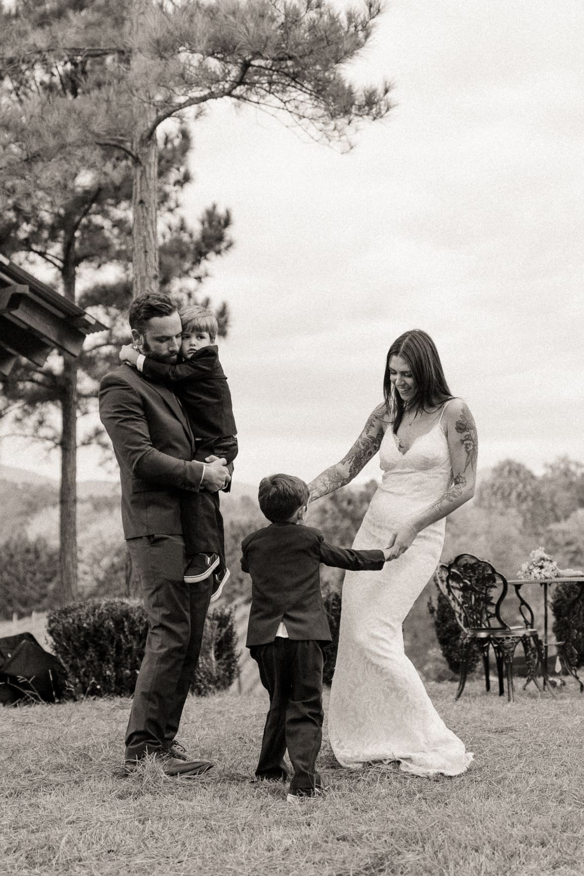 Groom holds a toddler while a ring bearer reaches for the bride's hand on a lawn beneath a tall pine