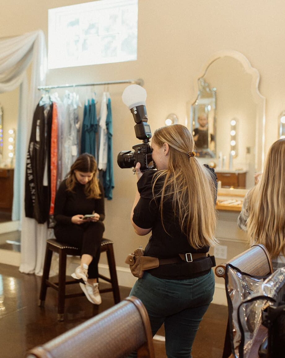 Wedding photographer works in a getting-ready room beside a clothing rack and vanity mirrors