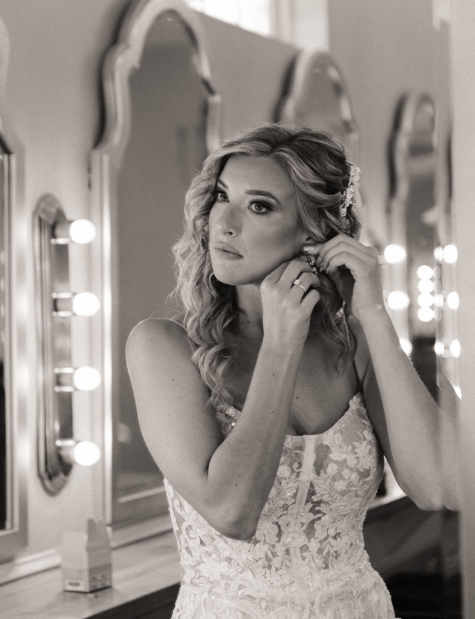 Bride in a lace gown puts on an earring in front of a lit vanity mirror, black and white wedding photo