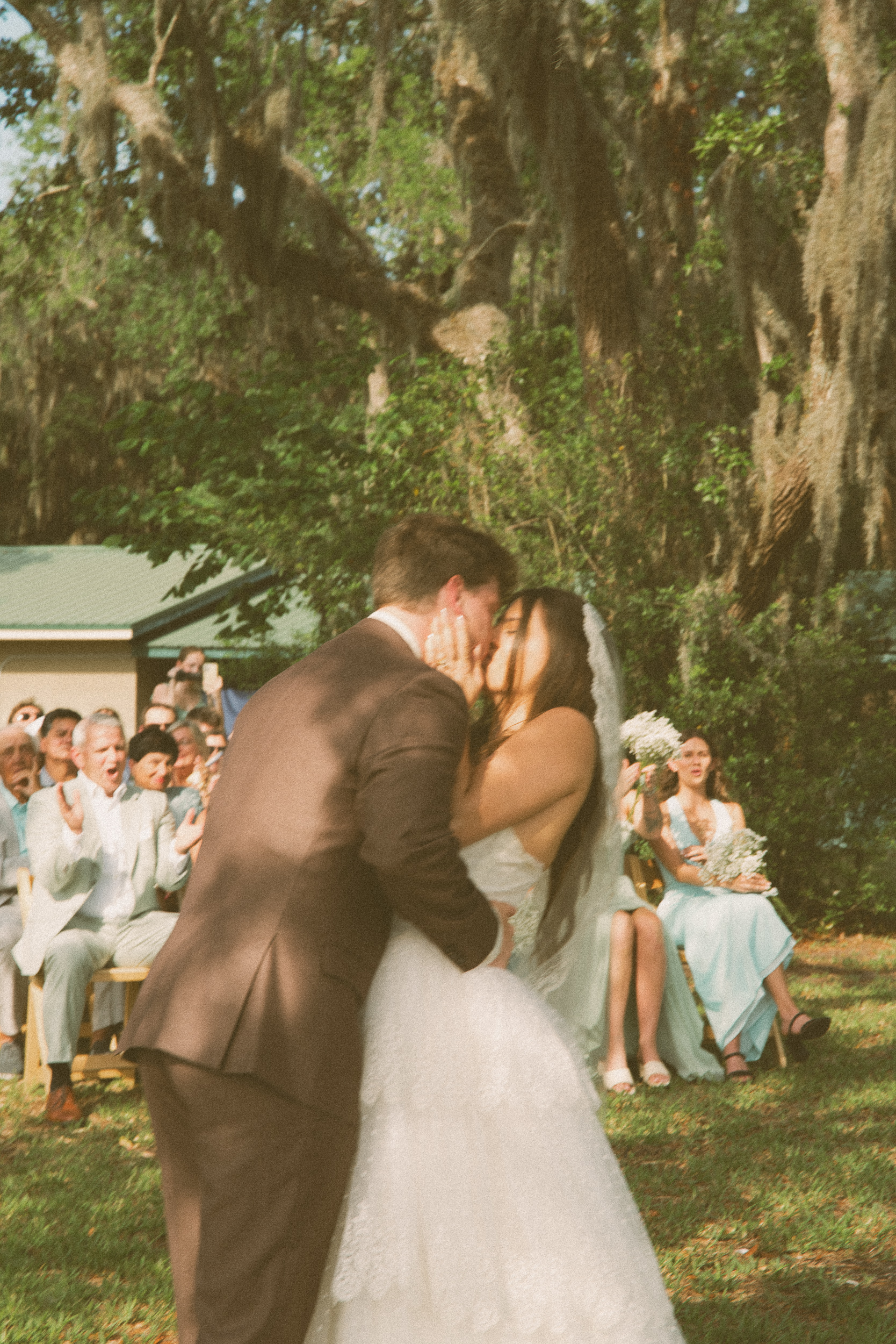 Couple's first kiss at an outdoor ceremony beneath moss-draped oaks as guests cheer behind them