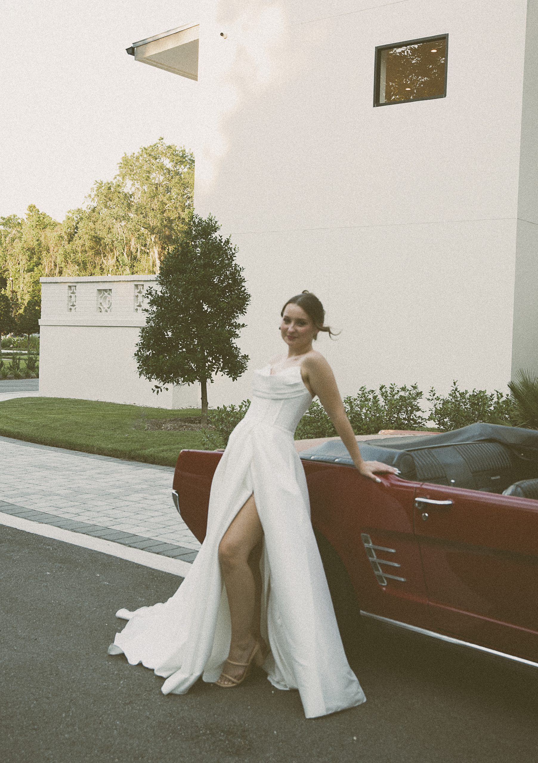 Bride in a high-slit gown leaning against a vintage red convertible in front of a modern white building