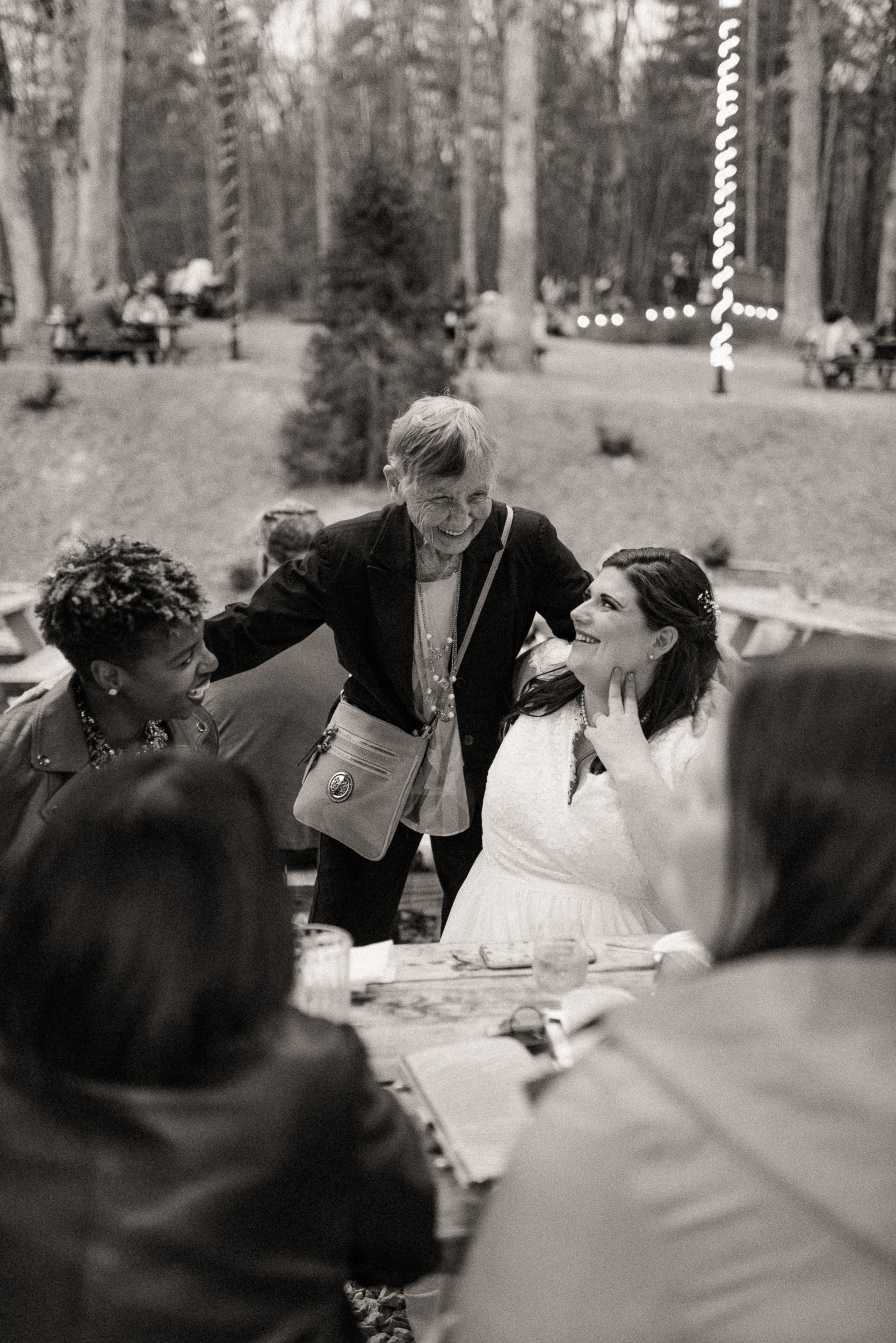 Black and white candid of a guest leaning in to hug the bride at a woodland reception table strung with lights