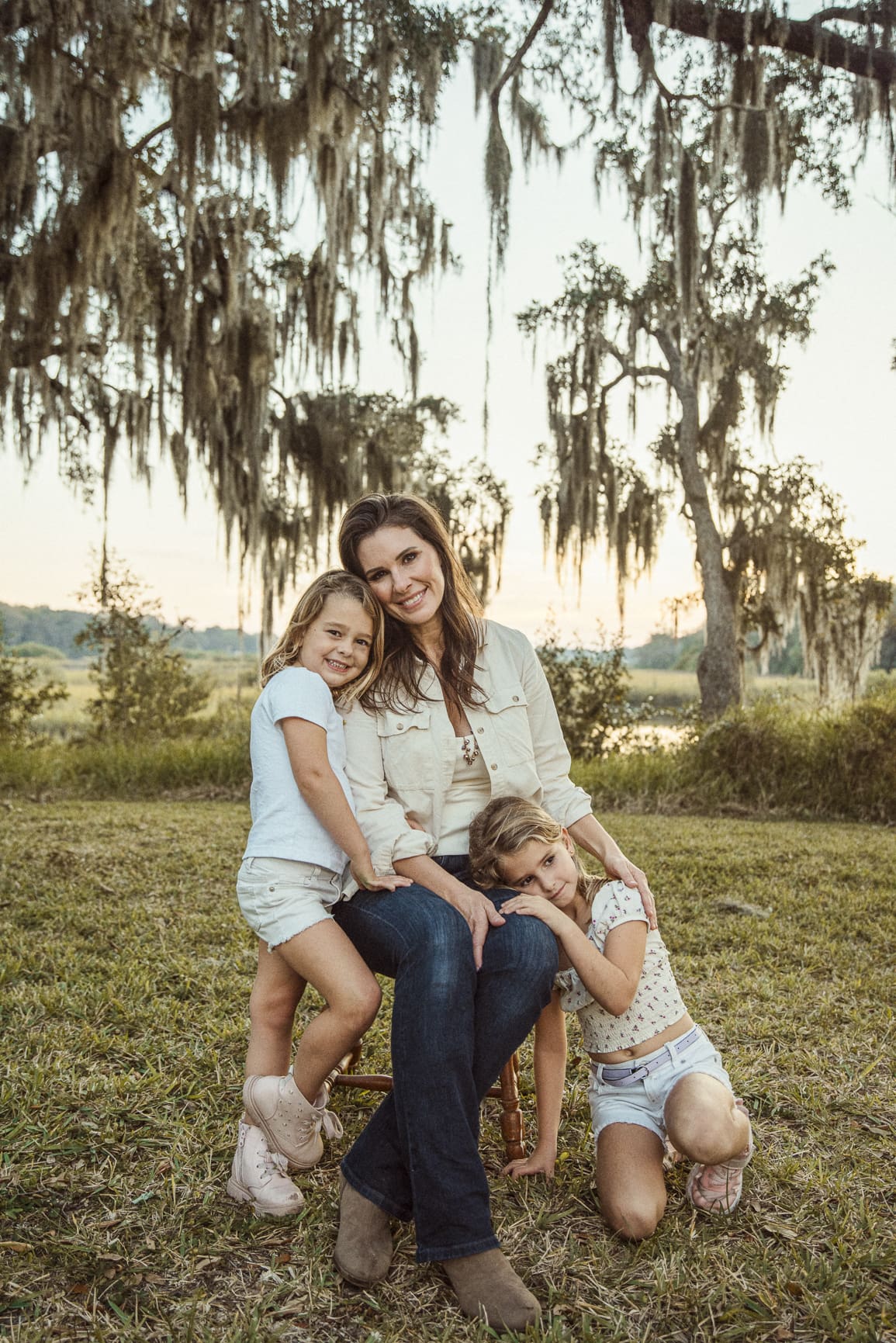 Mother and two daughters wearing soft neutral outfits during outdoor family photo session under oak trees. 