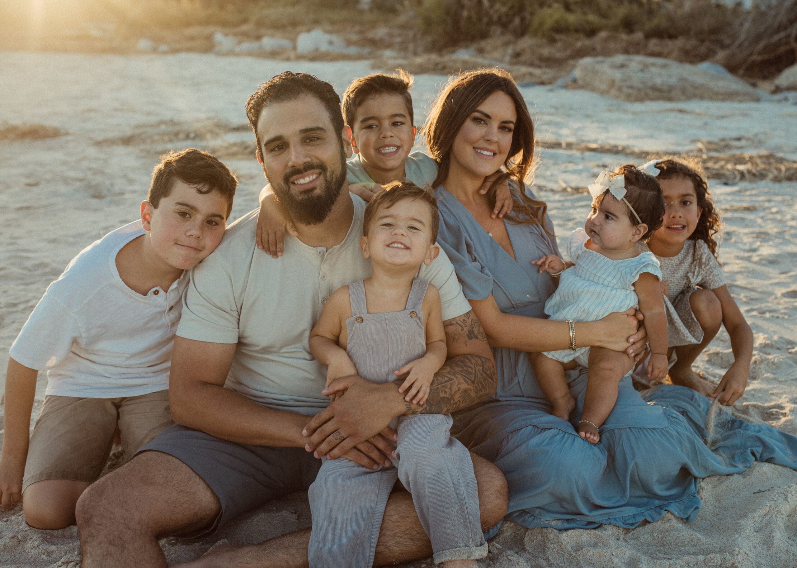 Family sitting together on beach wearing soft neutral outfits during golden hour session. 