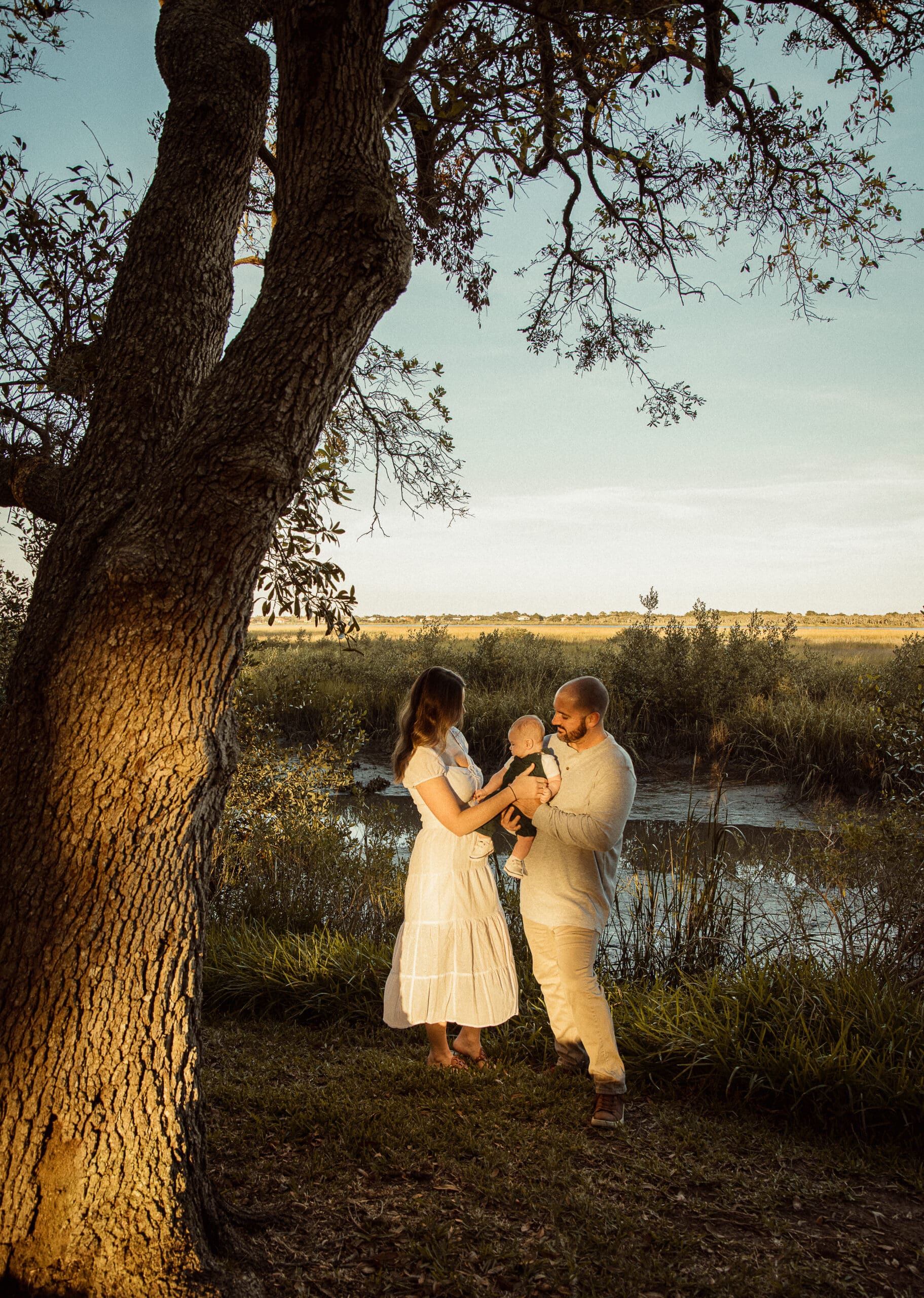 Family of three wearing light neutral outfits during golden hour by marsh water in St. Augustine