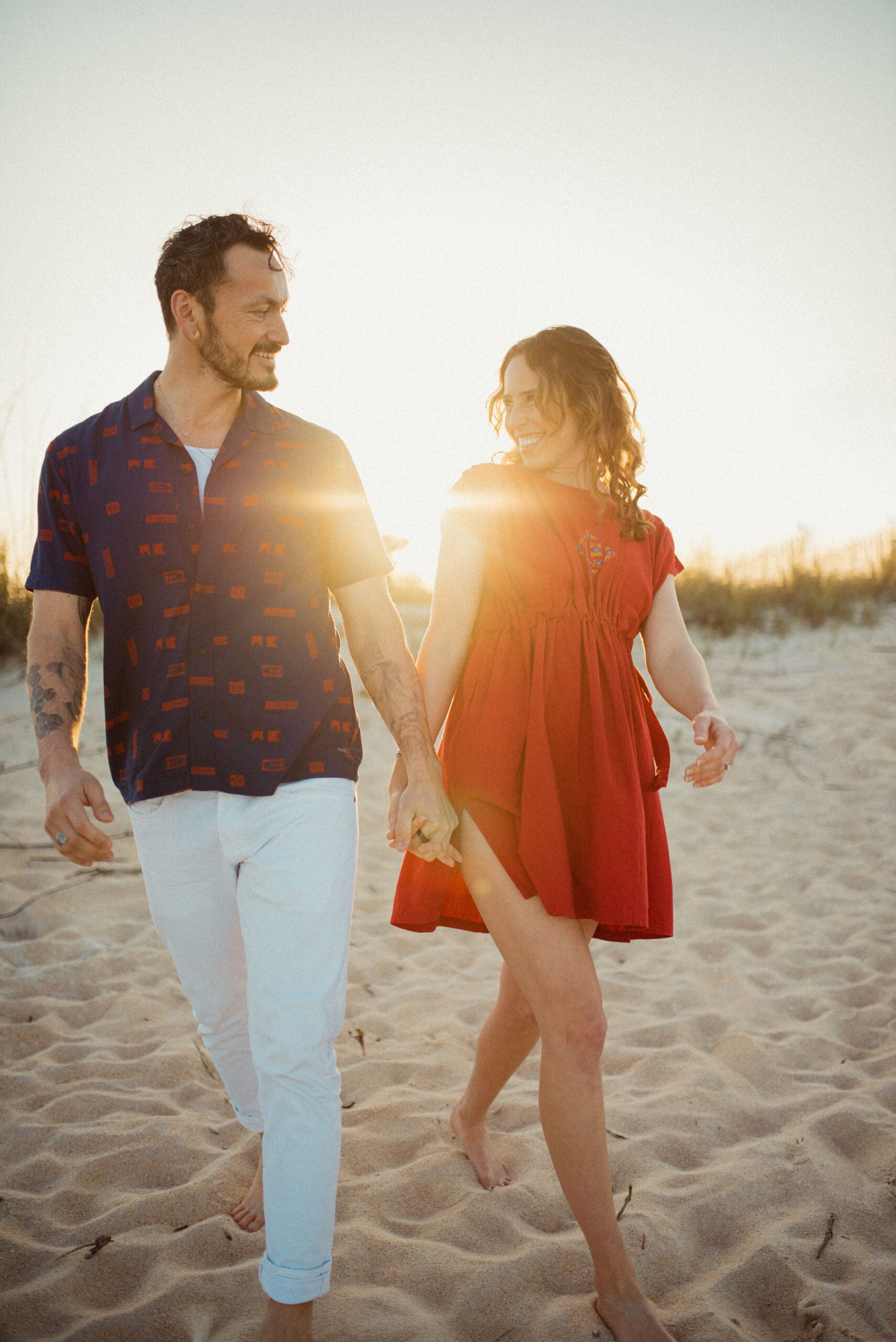 Couple walking on the beach at sunset wearing coordinated engagement photo outfits with a red dress and neutral tones
