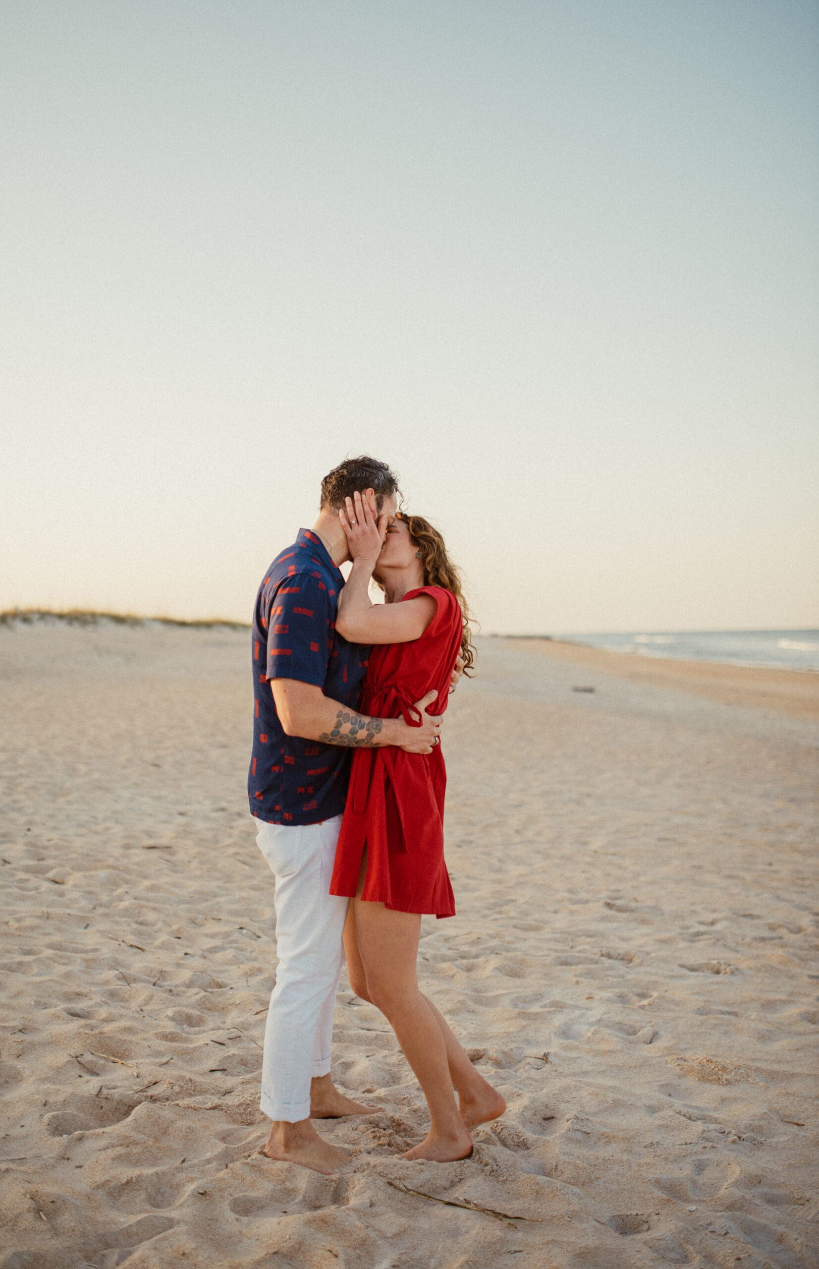 Couple kissing on the beach during engagement photos wearing a red dress and coordinated neutral outfit