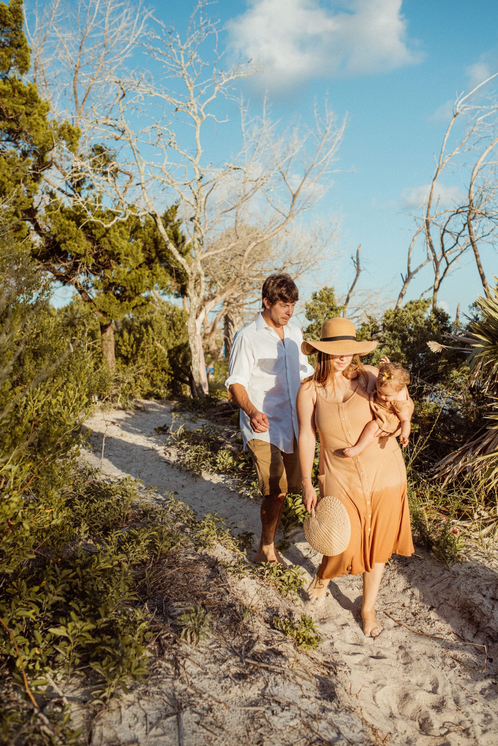 Family walkking on sandy path with mother in golden flowy dress and hat during sunset session. 