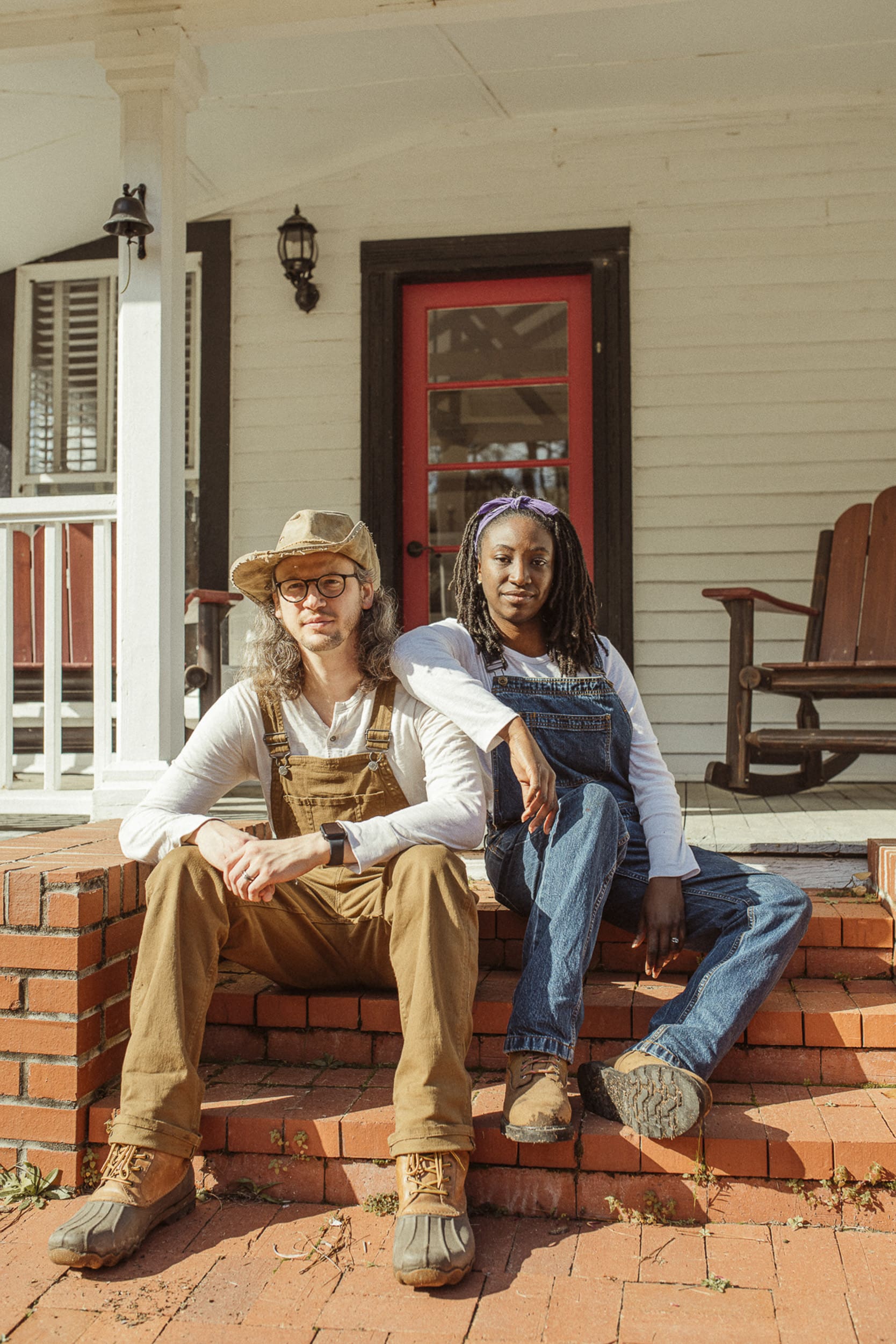 Couple wearing earthy toned outfits and overalls for engagement photos on a front porch