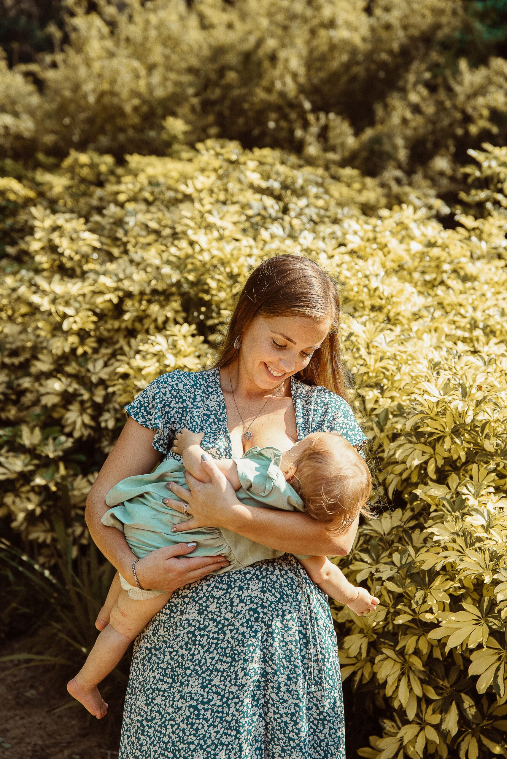 Mother holding baby in vintage green textured dress during outdoor family photo session with soft natural light. 