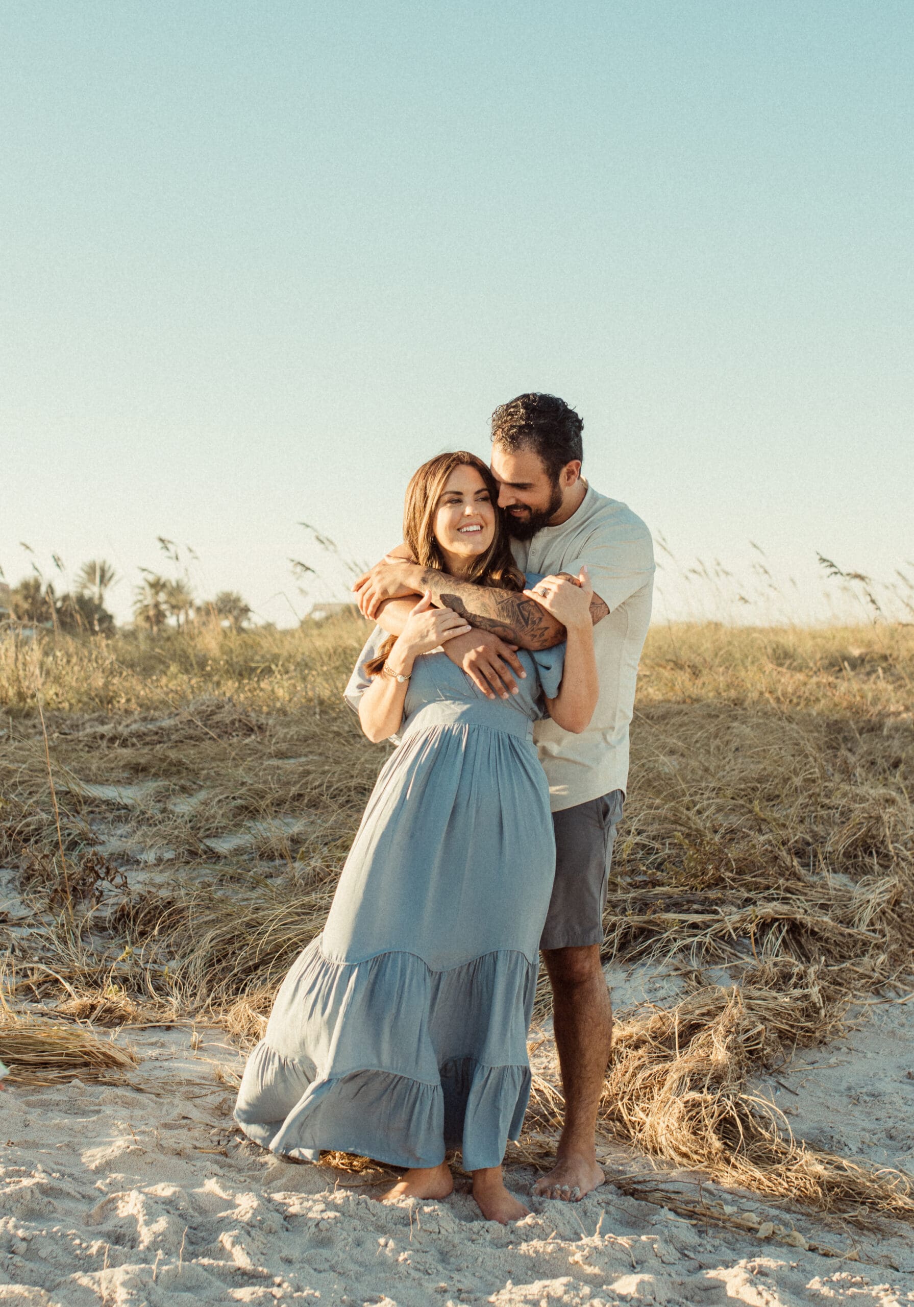 Couple standing near dunes at sunset hugging each other in soft flowing outfits during a romantic engagement session.