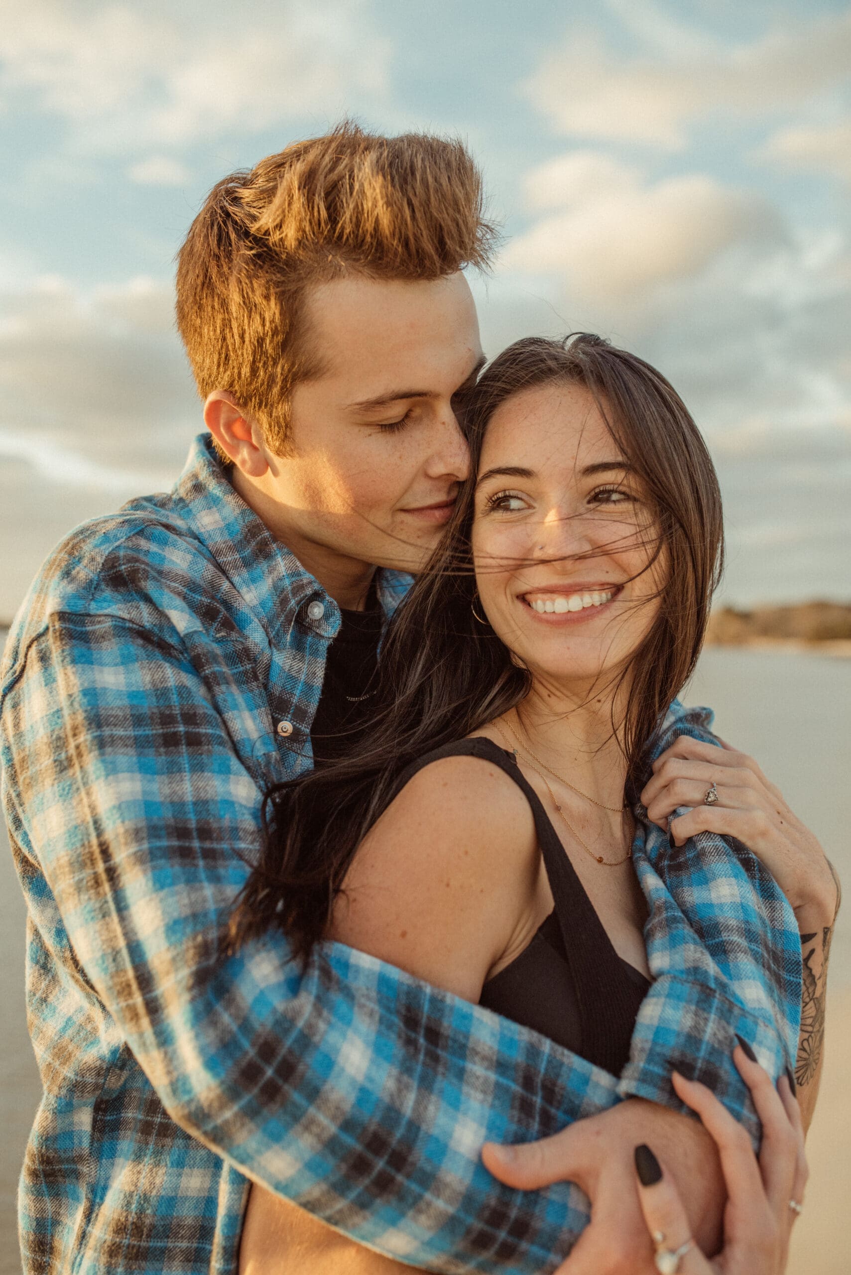 couple embracing on a windy beach capturing a candid and emotional romantic engagement moment. 