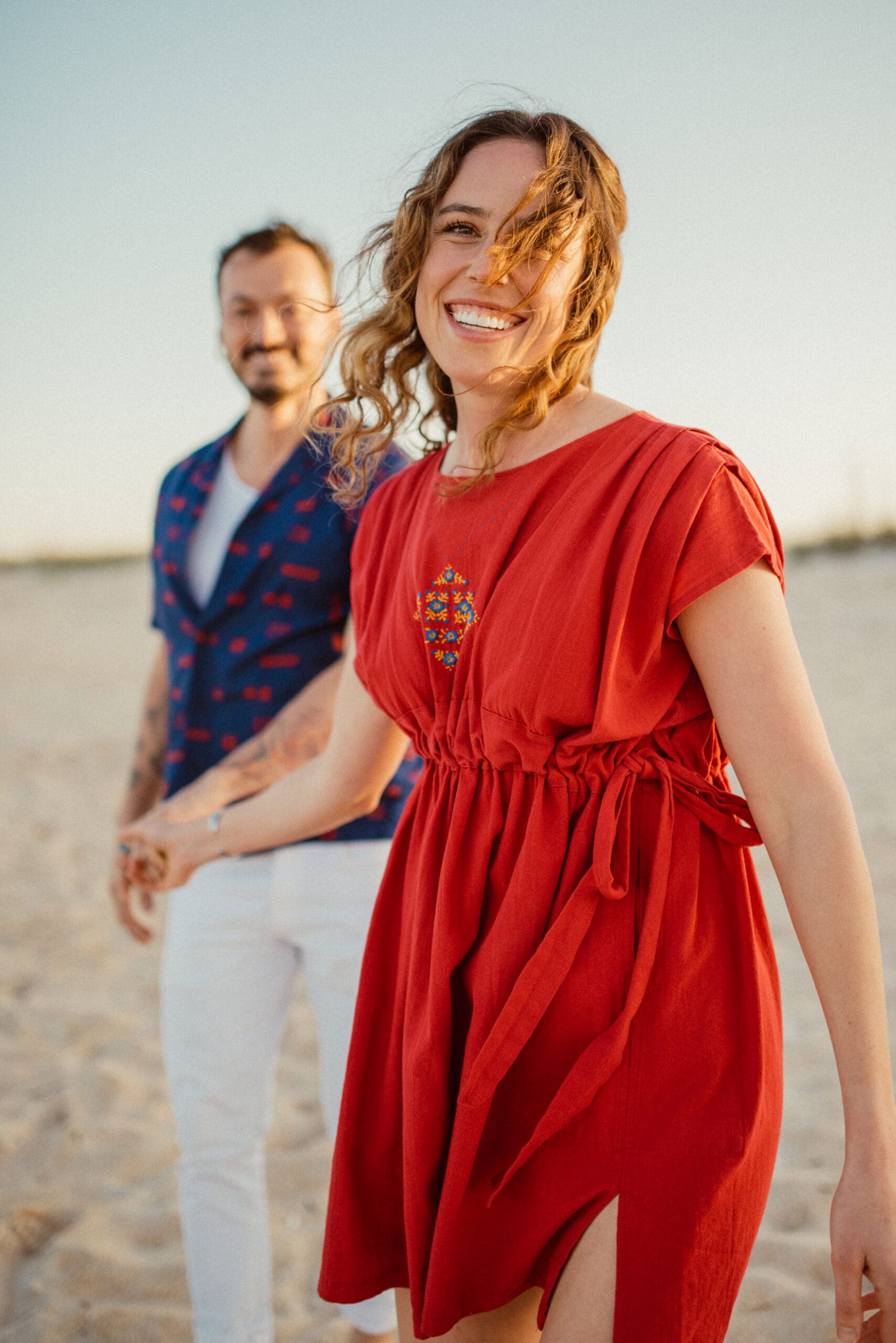 Woman in a flowing red dress smiling during a beach engagement session with soft movement and light